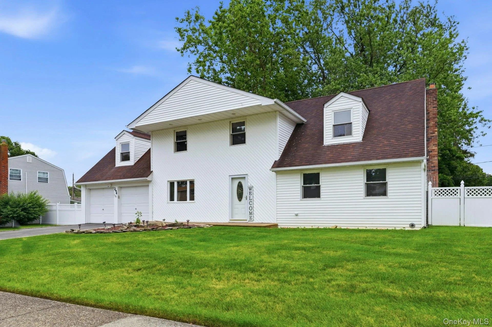 White siding exterior with a brown shingle roof, featuring a two-car garage, dormer window, front porch, and a white lattice fence White siding exterior with a brown shingle roof, featuring a two-car garage, dormer window, front porch, and a white lattice fence