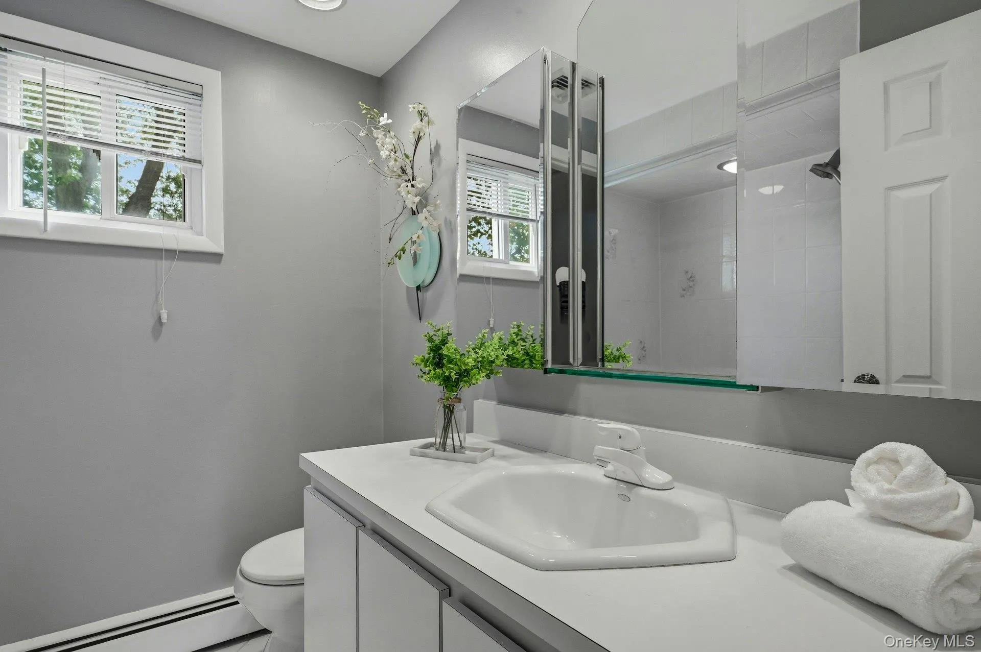 Bathroom featuring a white vanity with integrated sink, a medicine cabinet with mirrored doors, a window with blinds, and recessed lighting Bathroom featuring a white vanity with integrated sink, a medicine cabinet with mirrored doors, a window with blinds, and recessed lighting