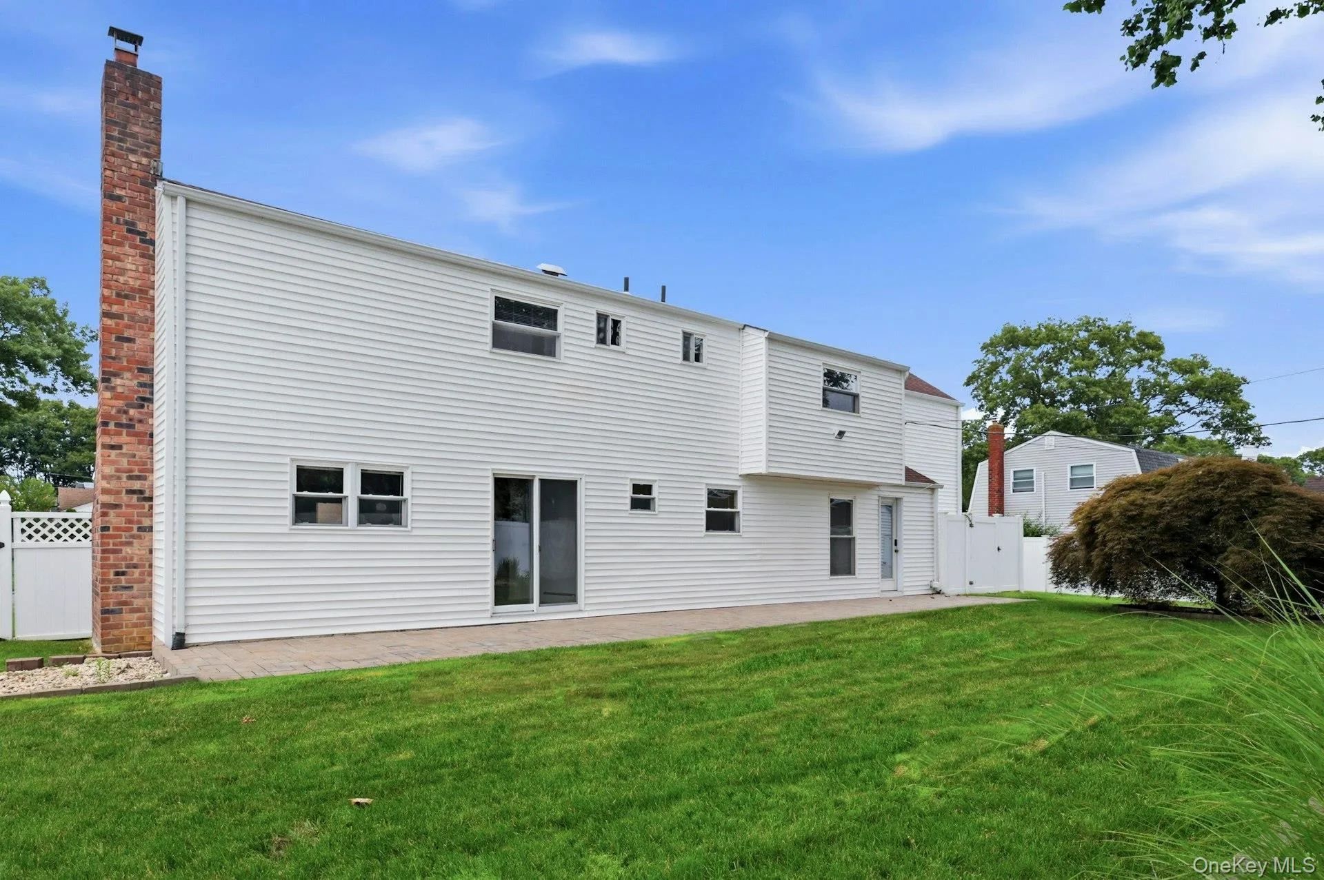 Rear exterior featuring white horizontal siding, a brick chimney, a ground-level patio, and a manicured lawn Rear exterior featuring white horizontal siding, a brick chimney, a ground-level patio, and a manicured lawn