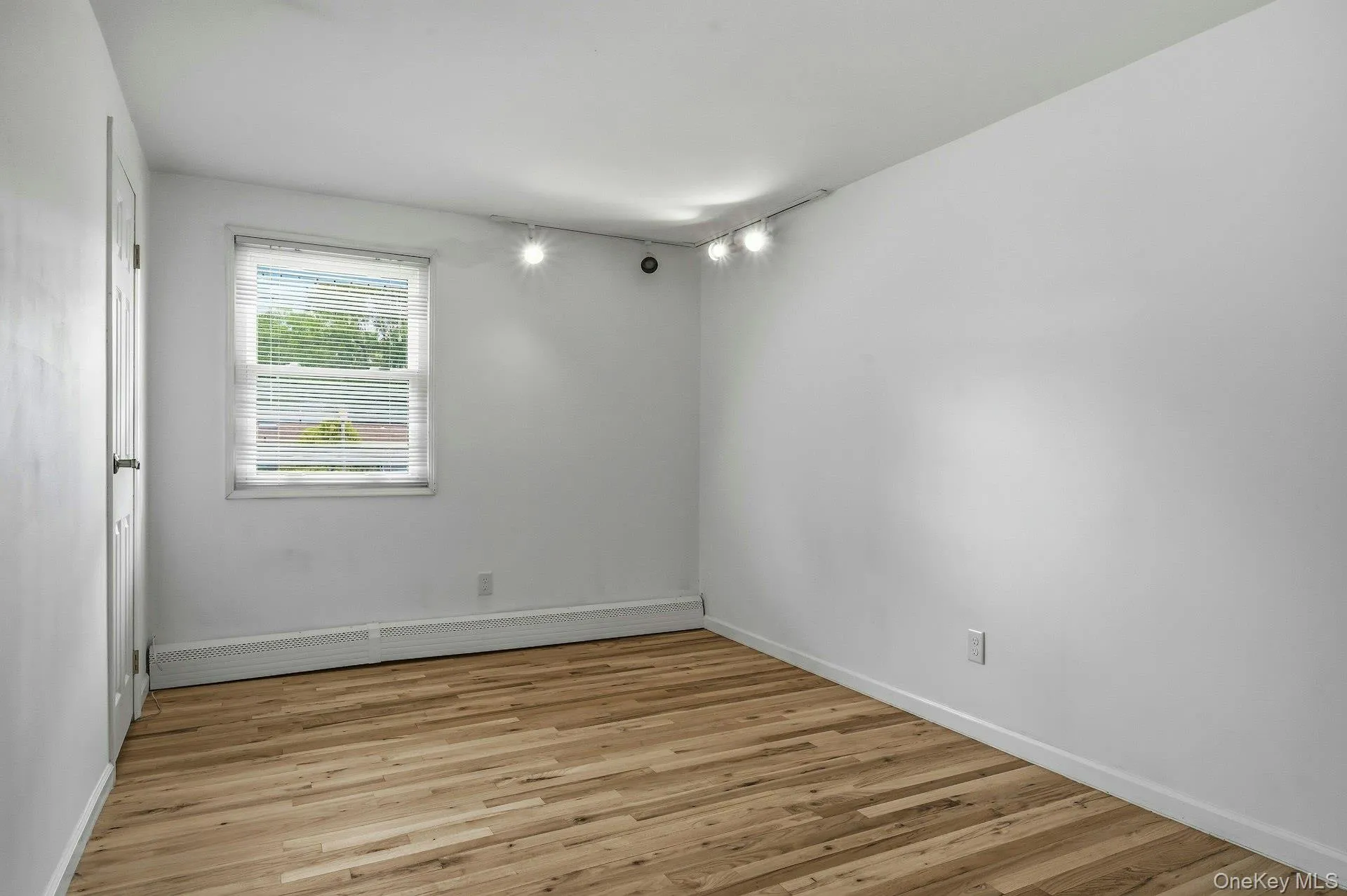 Room featuring wood-finish flooring, white painted walls, a single window with blinds, a baseboard heater, and ceiling-mounted track lighting Room featuring wood-finish flooring, white painted walls, a single window with blinds, a baseboard heater, and ceiling-mounted track lighting