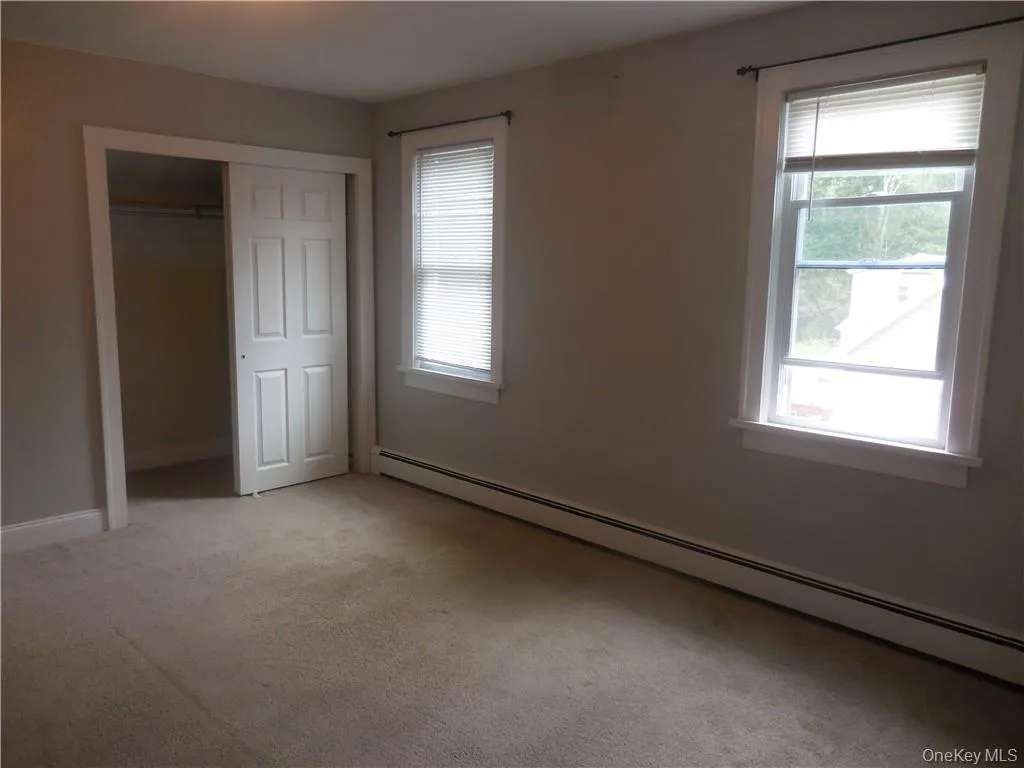 Unfurnished bedroom with a closet, multiple windows, baseboard heating, and light carpet Unfurnished bedroom with a closet, multiple windows, baseboard heating, and light carpet