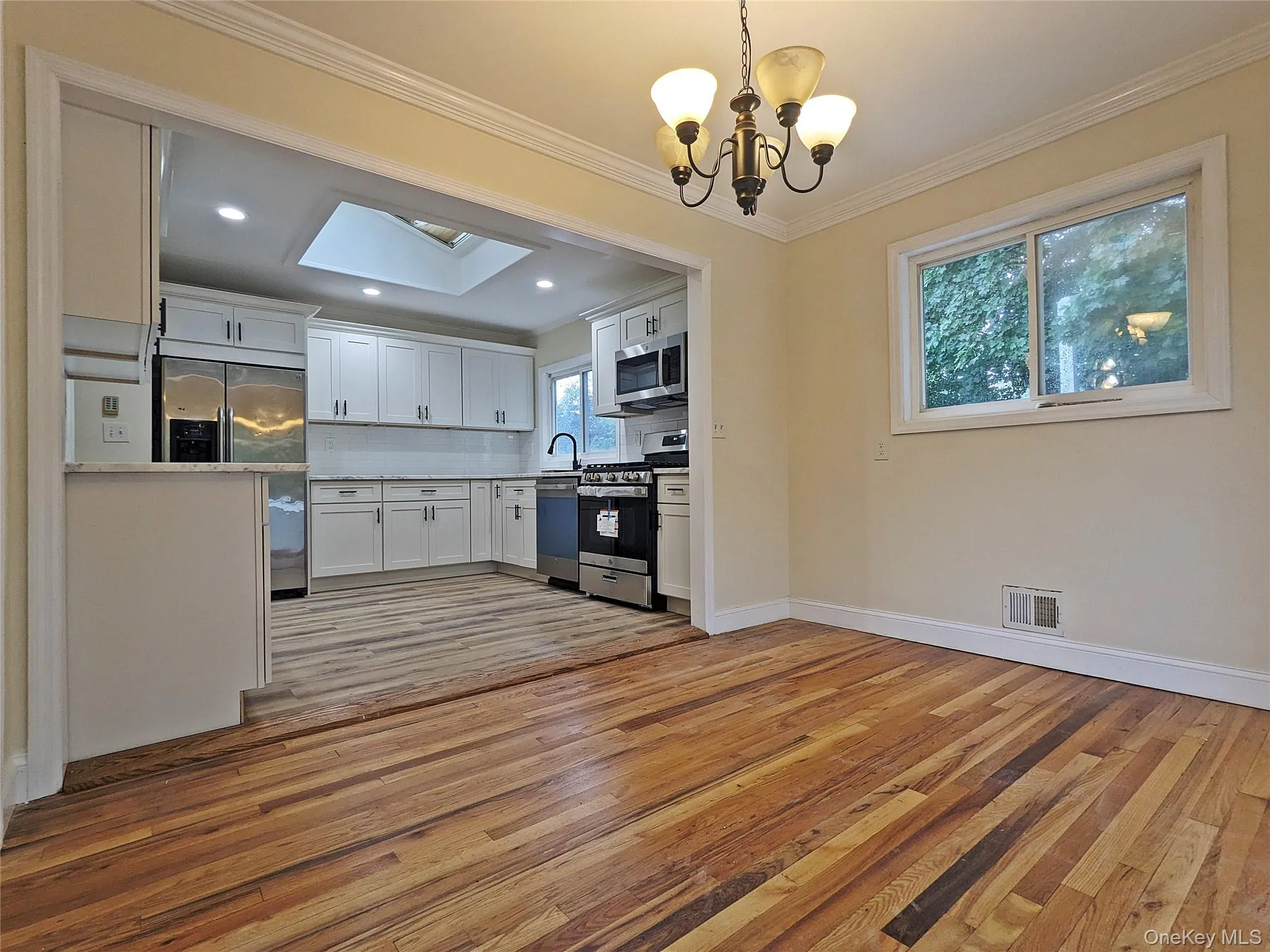Open-concept dining area with wood-finish flooring, crown molding, and a chandelier, opening to a kitchen featuring white cabinetry, a skylight, and stainless steel appliances Open-concept dining area with wood-finish flooring, crown molding, and a chandelier, opening to a kitchen featuring white cabinetry, a skylight, and stainless steel appliances