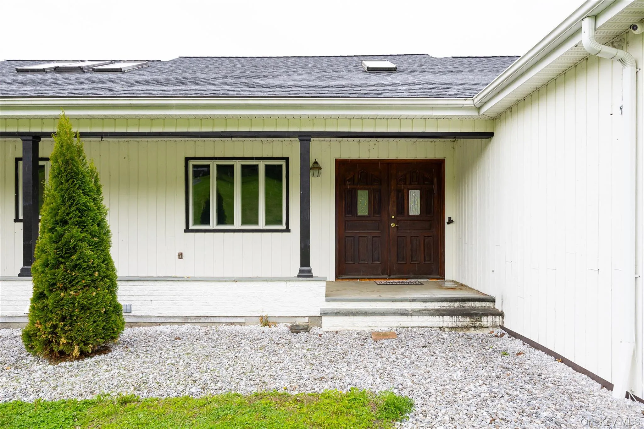 Front entry featuring double dark wood doors with glass inserts, white vertical siding, black support columns, and a dark shingle roof with skylights Front entry featuring double dark wood doors with glass inserts, white vertical siding, black support columns, and a dark shingle roof with skylights