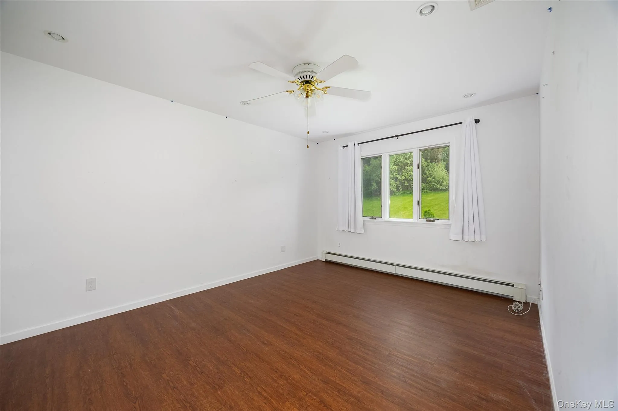 Room featuring wood-finish flooring, white walls, and a multi-pane window with a green outdoor scenery Room featuring wood-finish flooring, white walls, and a multi-pane window with a green outdoor scenery