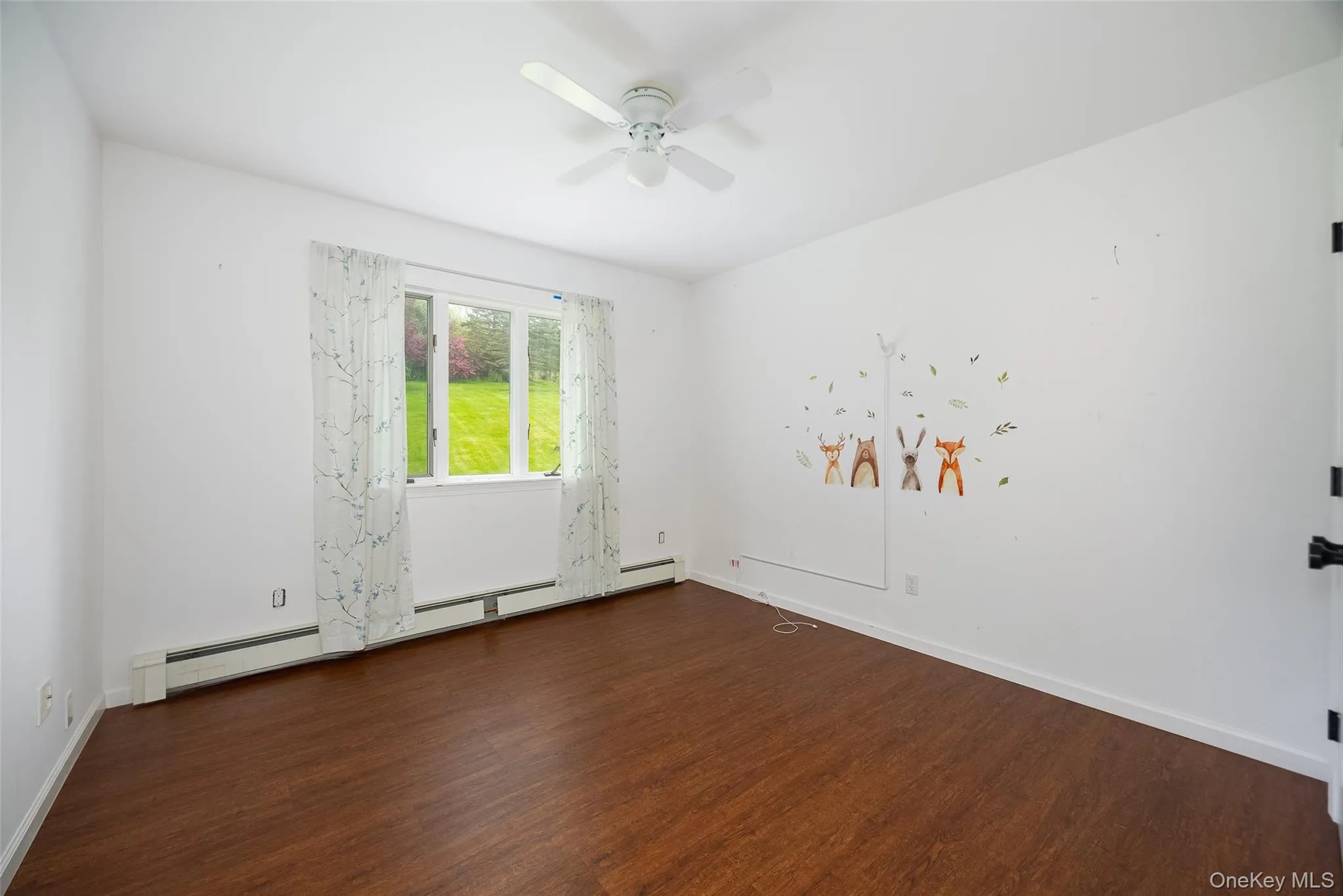 Bright interior space featuring wood-finish flooring, a ceiling fan, and a large window overlooking a verdant outdoor area Bright interior space featuring wood-finish flooring, a ceiling fan, and a large window overlooking a verdant outdoor area