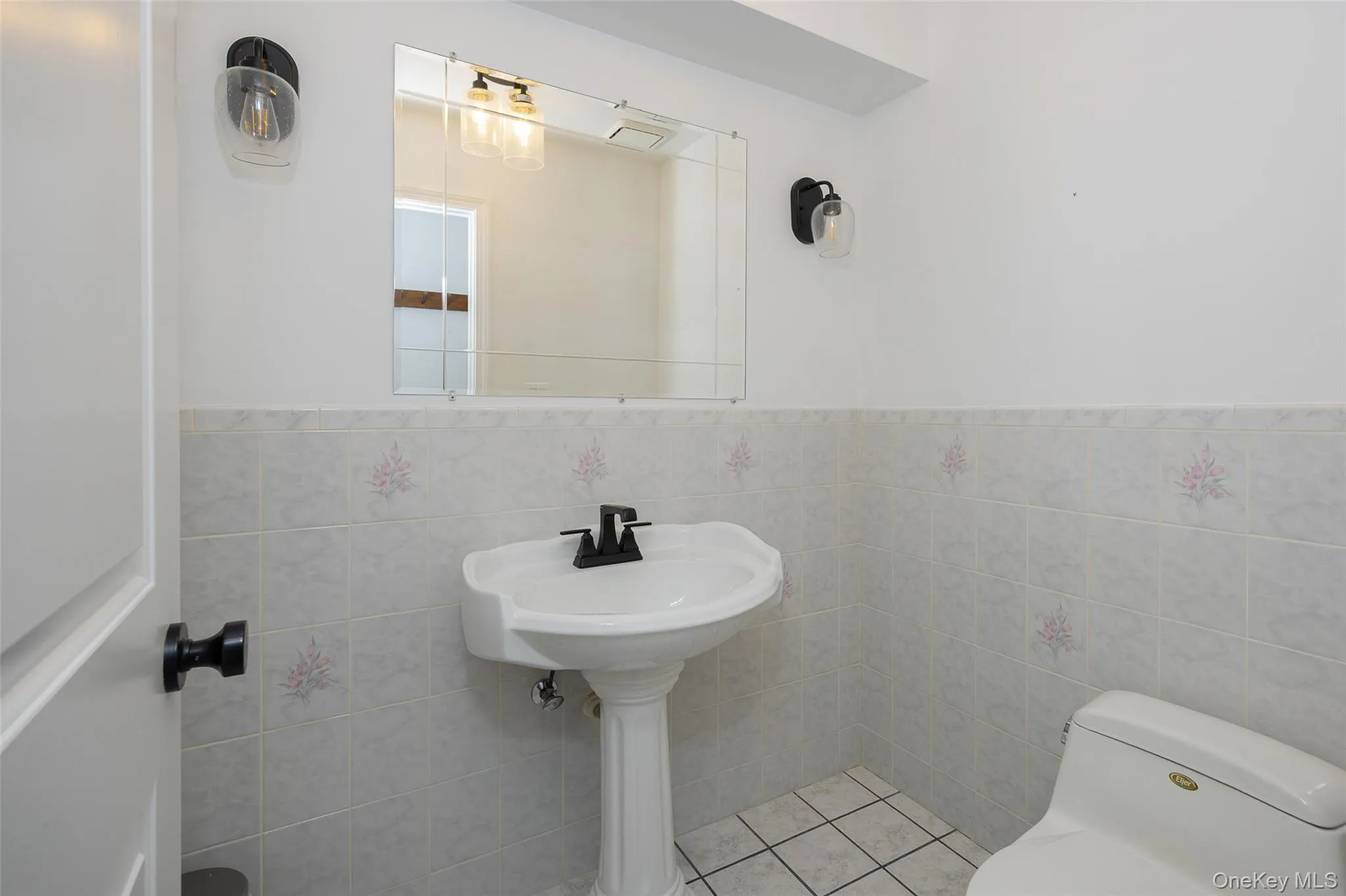 Pedestal sink with a black faucet, white tiled walls with subtle floral accents, and a complementary white toilet Pedestal sink with a black faucet, white tiled walls with subtle floral accents, and a complementary white toilet