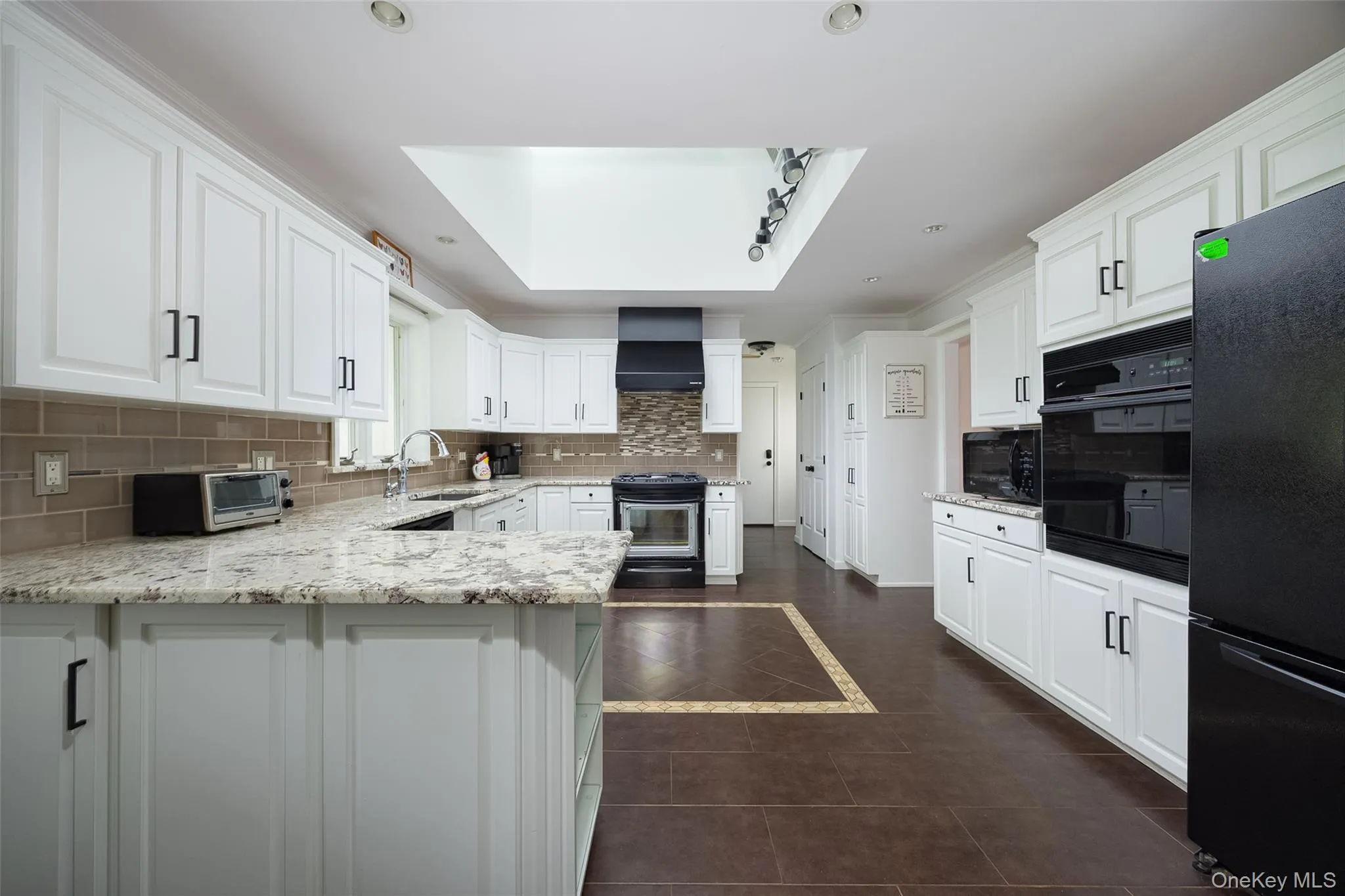 Kitchen featuring white cabinetry, light-toned countertops, a brown subway tile backsplash, dark-finish appliances, and a skylight Kitchen featuring white cabinetry, light-toned countertops, a brown subway tile backsplash, dark-finish appliances, and a skylight