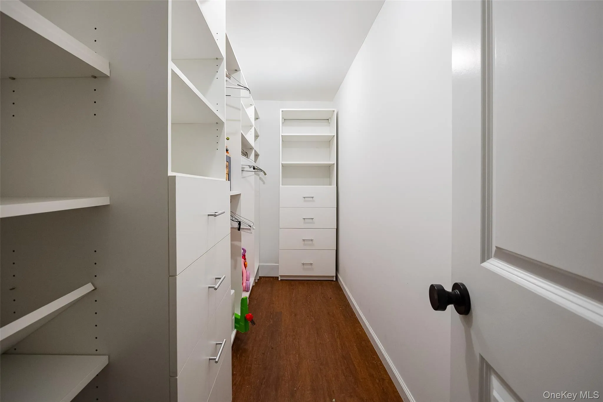Walk-in closet featuring built-in white shelving, integrated drawers with metal pulls, ample hanging space, and wood-finish flooring Walk-in closet featuring built-in white shelving, integrated drawers with metal pulls, ample hanging space, and wood-finish flooring