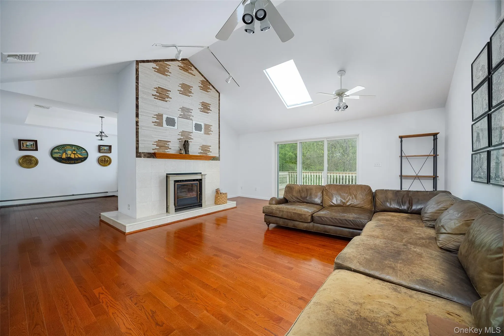 Spacious living area featuring polished wood-finish flooring, a prominent fireplace with a decorative tile surround, vaulted ceilings, and a large skylight Spacious living area featuring polished wood-finish flooring, a prominent fireplace with a decorative tile surround, vaulted ceilings, and a large skylight
