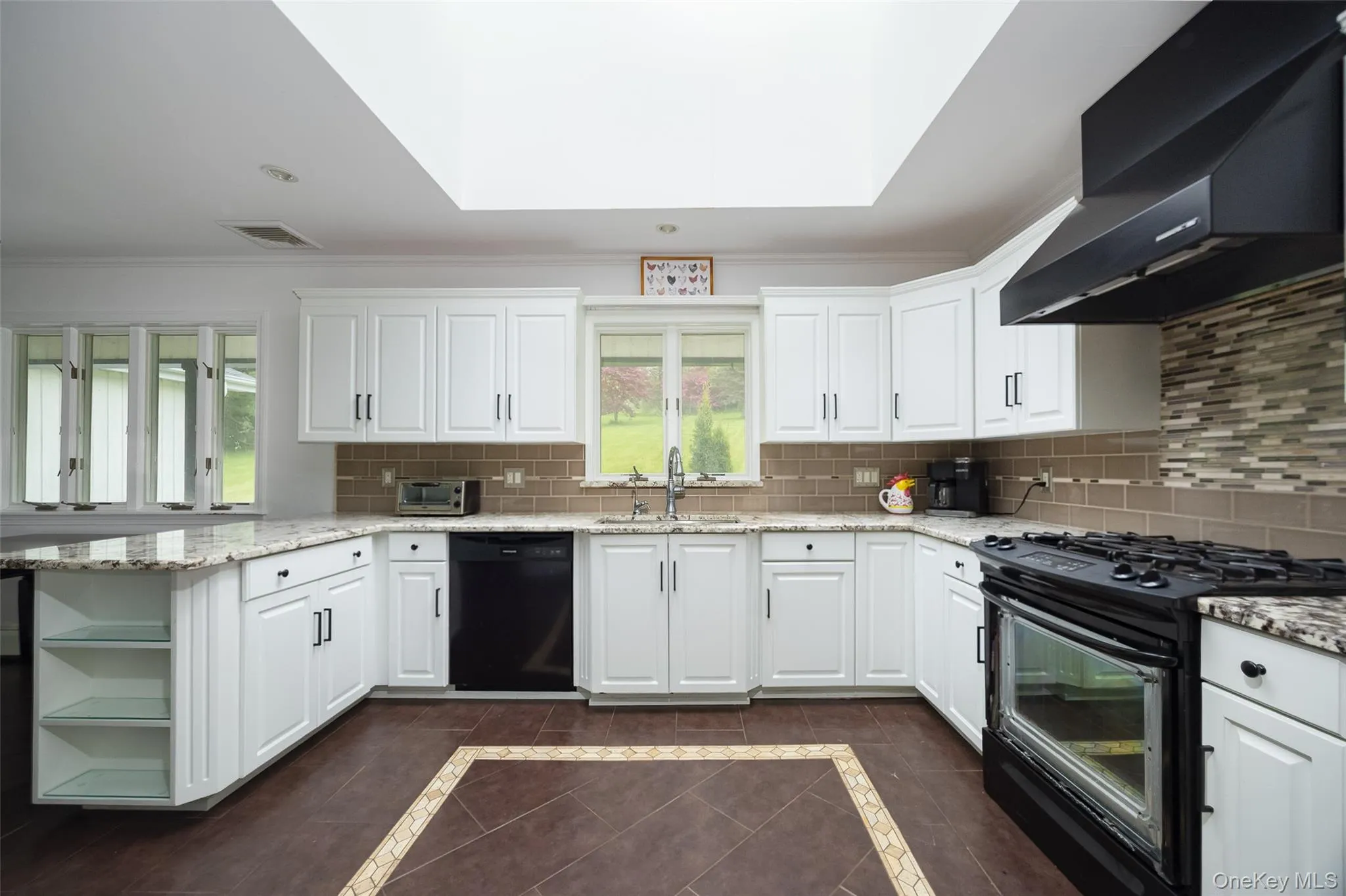 Bright kitchen featuring white cabinetry, stone countertops, a subway tile backsplash, a black gas range with coordinating hood, and a skylight Bright kitchen featuring white cabinetry, stone countertops, a subway tile backsplash, a black gas range with coordinating hood, and a skylight