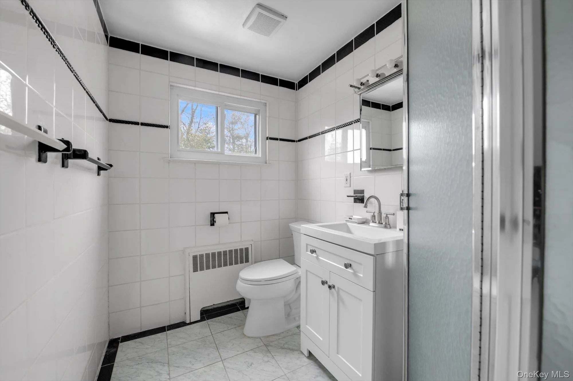 Bathroom featuring white subway tile walls with black trim, a white vanity with cabinet storage, a wall-mounted mirror, and a window providing natural light Bathroom featuring white subway tile walls with black trim, a white vanity with cabinet storage, a wall-mounted mirror, and a window providing natural light