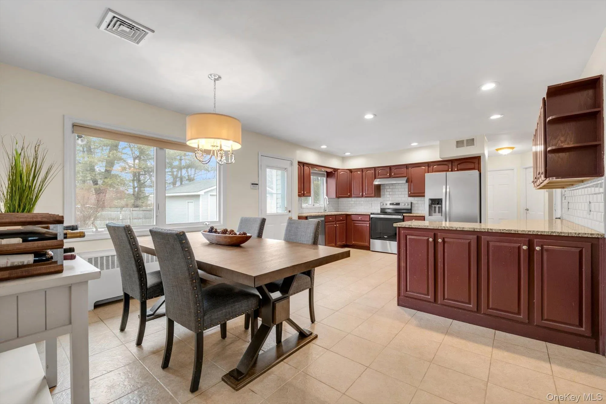 Open-concept kitchen and dining area featuring wood-finish cabinetry, stainless steel appliances, tile flooring, recessed lighting, and a large window Open-concept kitchen and dining area featuring wood-finish cabinetry, stainless steel appliances, tile flooring, recessed lighting, and a large window