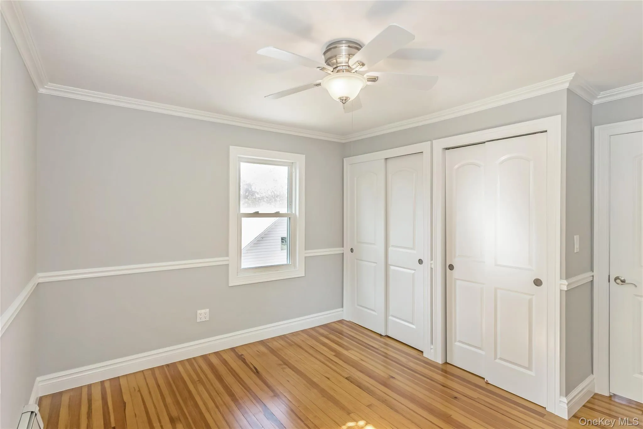 Unfurnished bedroom featuring two closets, light wood finished floors, ornamental molding, ceiling fan, and a baseboard radiator Unfurnished bedroom featuring two closets, light wood finished floors, ornamental molding, ceiling fan, and a baseboard radiator
