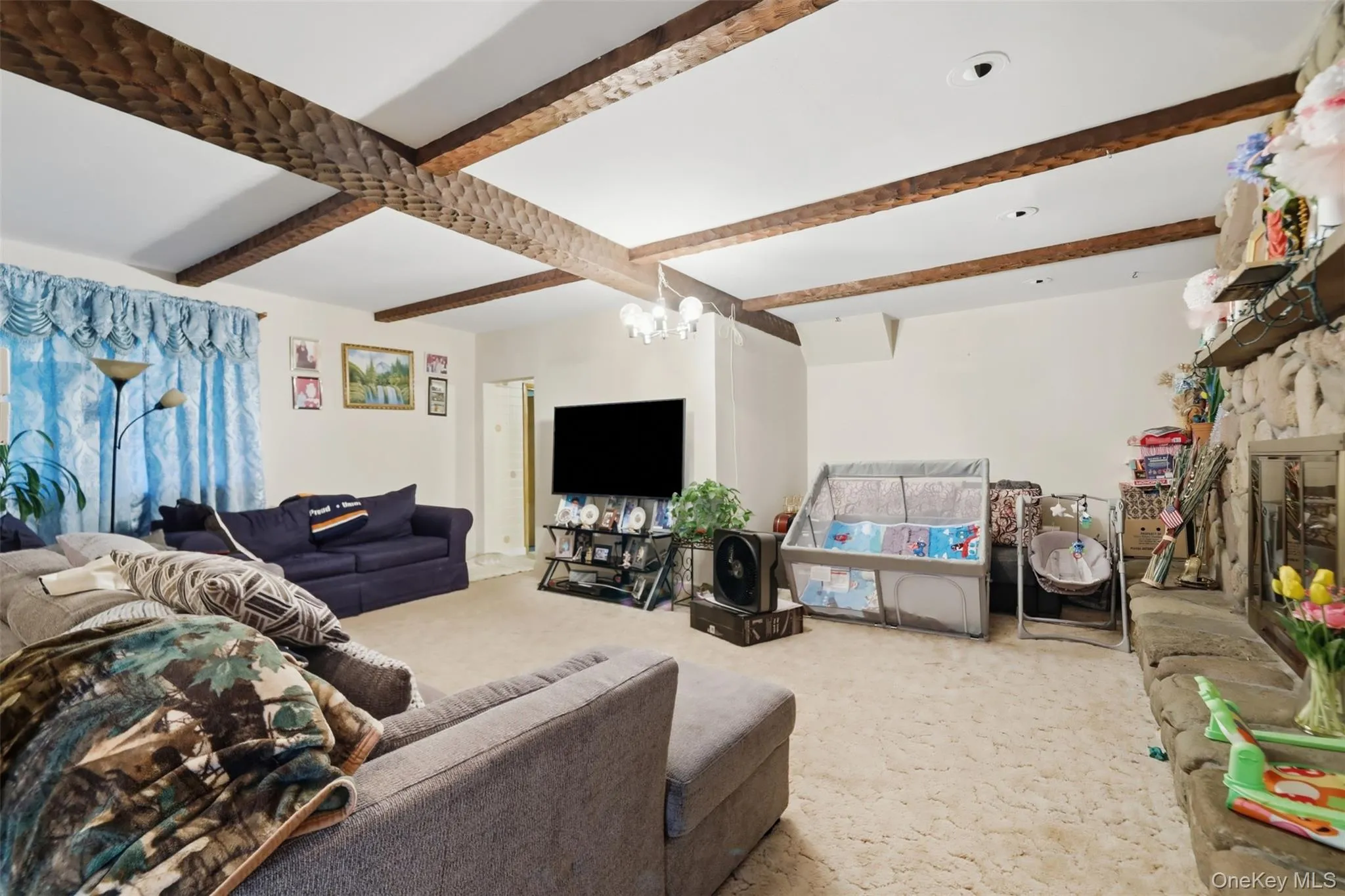 Living area featuring textured wood ceiling beams, a stone-clad fireplace with a hearth, recessed lighting, and light-toned carpeting Living area featuring textured wood ceiling beams, a stone-clad fireplace with a hearth, recessed lighting, and light-toned carpeting