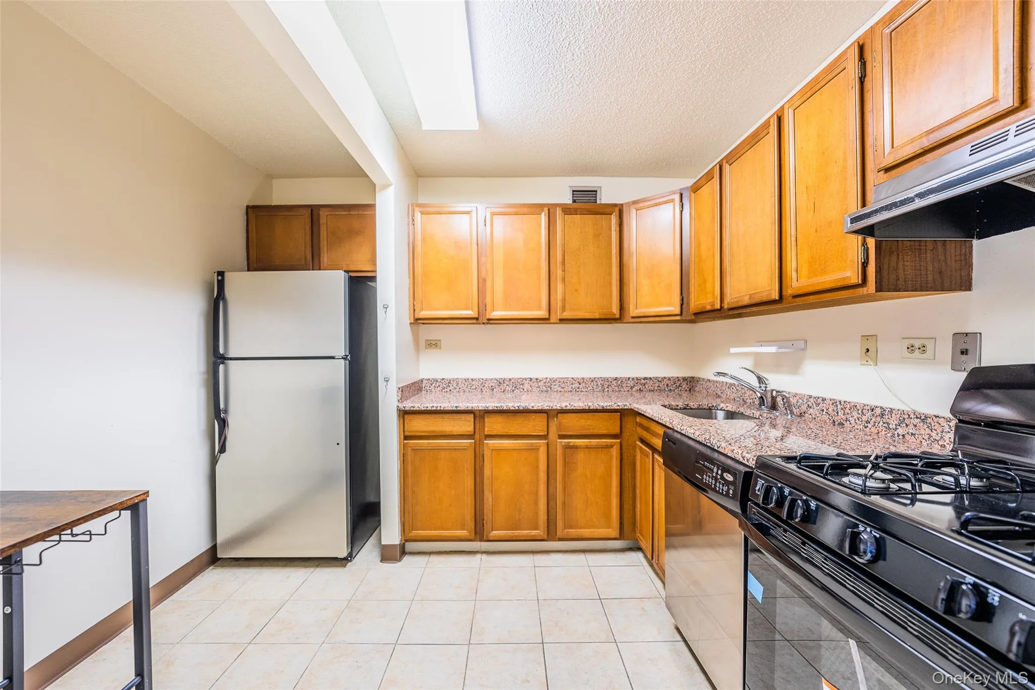Kitchen featuring stainless steel appliances, light stone counters, brown cabinets, a textured ceiling, and light tile patterned floors Kitchen featuring stainless steel appliances, light stone counters, brown cabinets, a textured ceiling, and light tile patterned floors