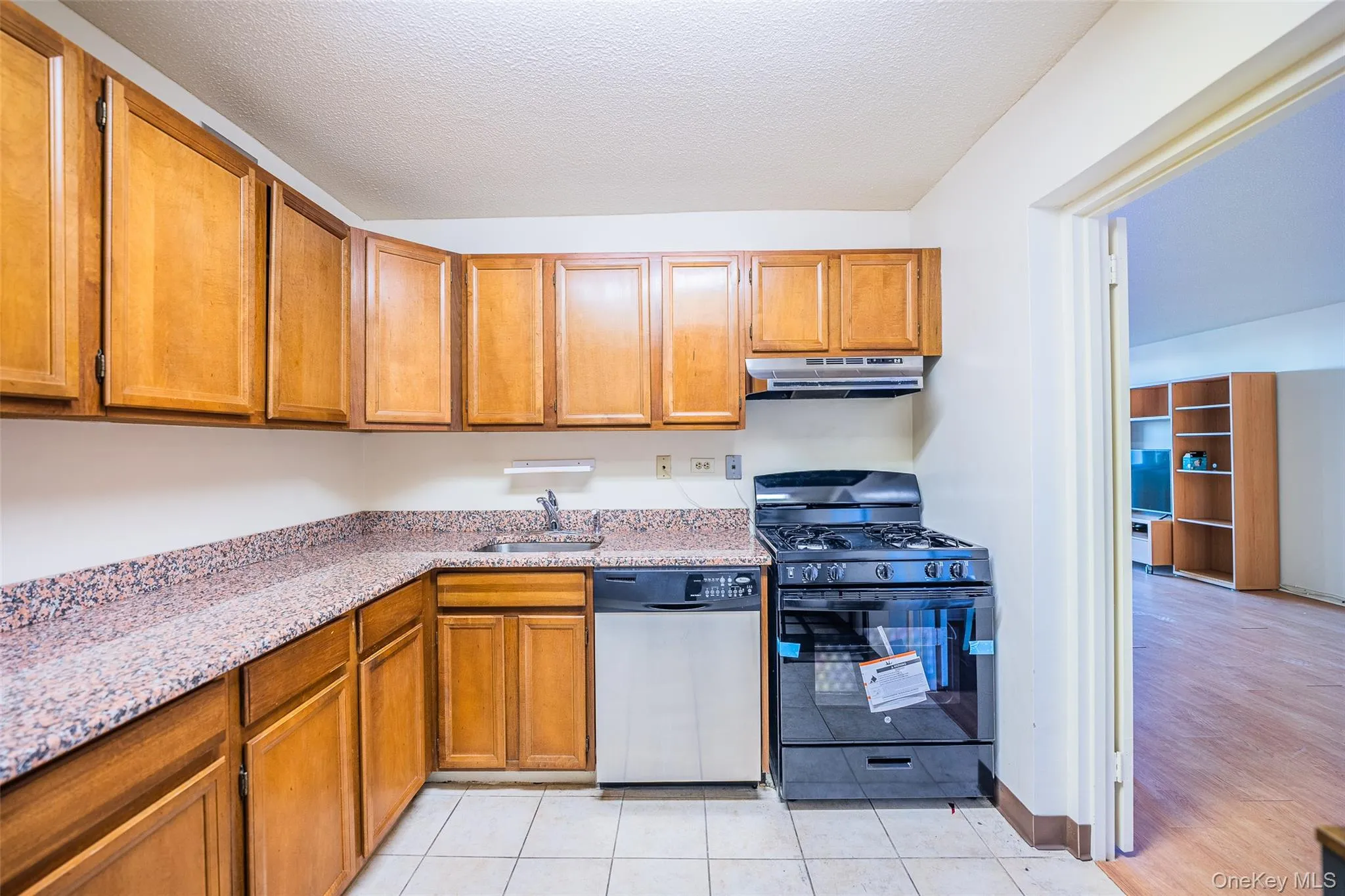 Kitchen featuring black gas range, stainless steel dishwasher, brown cabinetry, light stone counters, and under cabinet range hood Kitchen featuring black gas range, stainless steel dishwasher, brown cabinetry, light stone counters, and under cabinet range hood