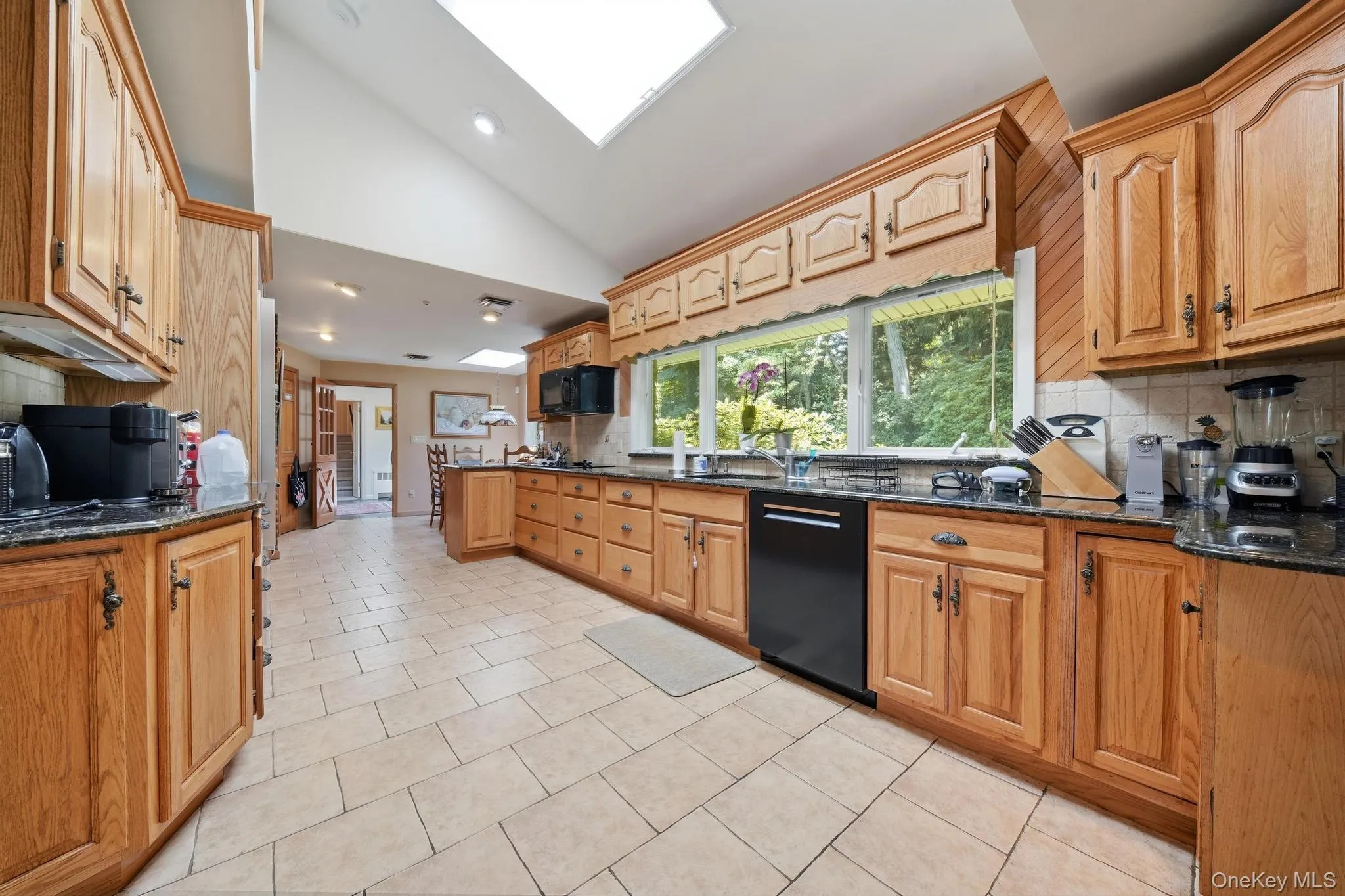 Kitchen featuring abundant wood cabinetry, dark countertops, a black dishwasher, and a large window overlooking trees Kitchen featuring abundant wood cabinetry, dark countertops, a black dishwasher, and a large window overlooking trees