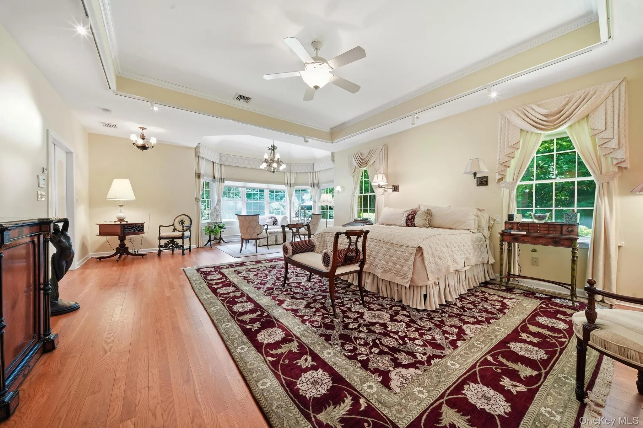 Spacious bedroom featuring wood-finish flooring, a tray ceiling with a ceiling fan, track lighting, and a bay window area Spacious bedroom featuring wood-finish flooring, a tray ceiling with a ceiling fan, track lighting, and a bay window area