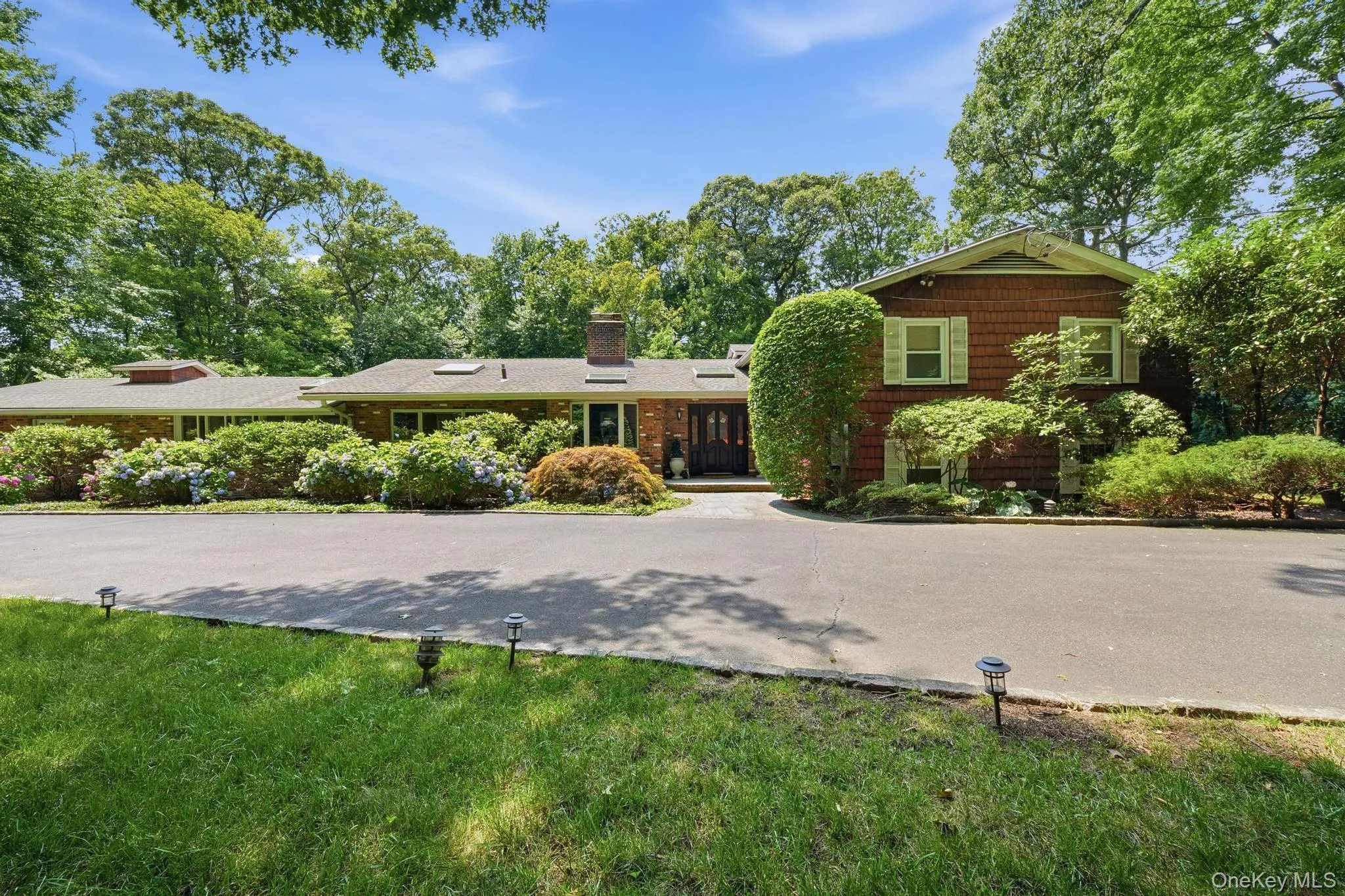 Ranch-style residence featuring a brick facade with cedar shake siding, a multi-car driveway, and integrated outdoor lighting Ranch-style residence featuring a brick facade with cedar shake siding, a multi-car driveway, and integrated outdoor lighting