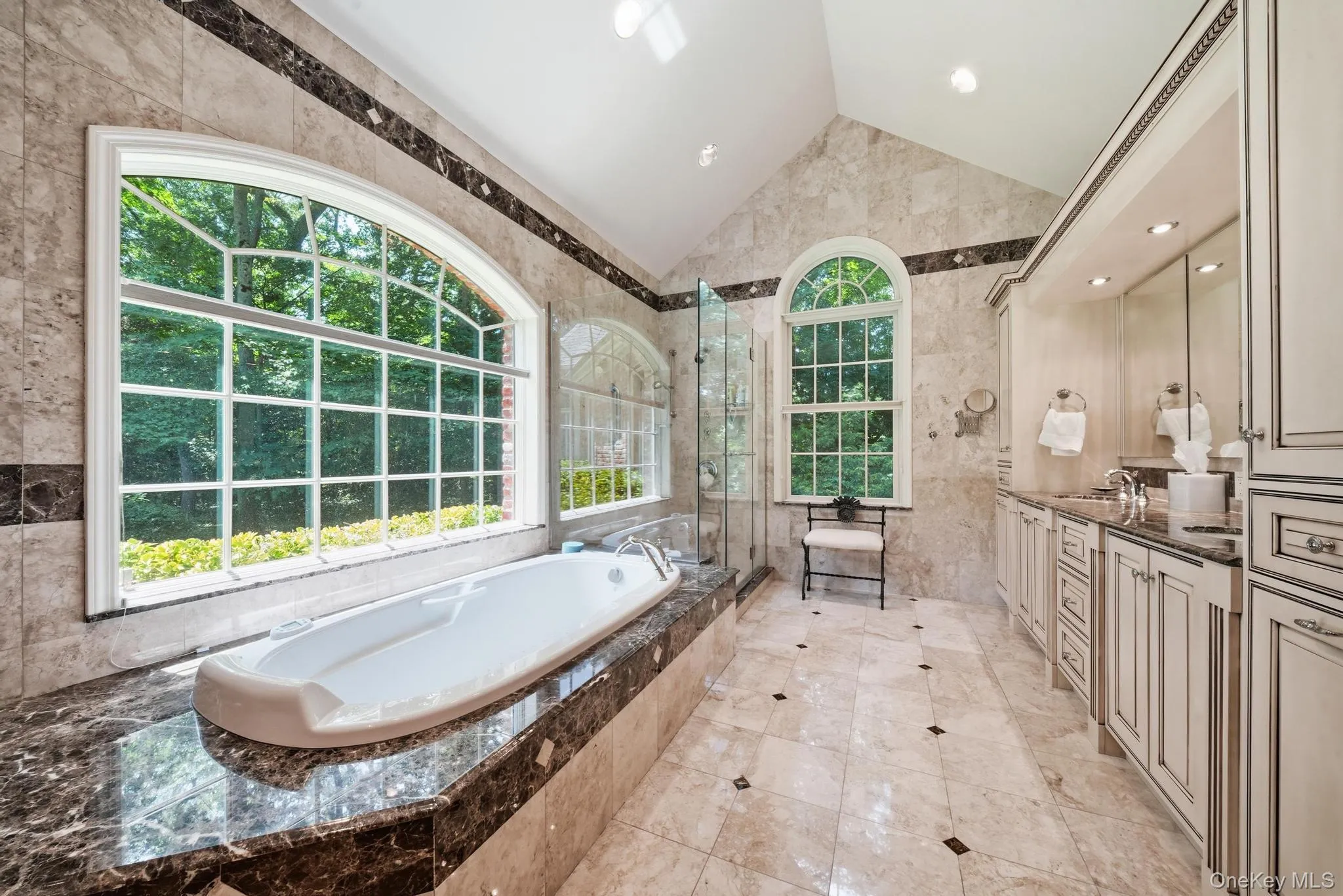 Luxurious bathroom featuring a large arched window, a built-in soaking tub with a dark stone surround, a separate glass-enclosed shower, a double vanity with dark countertops, and light tile flooring with dark accents Luxurious bathroom featuring a large arched window, a built-in soaking tub with a dark stone surround, a separate glass-enclosed shower, a double vanity with dark countertops, and light tile flooring with dark accents