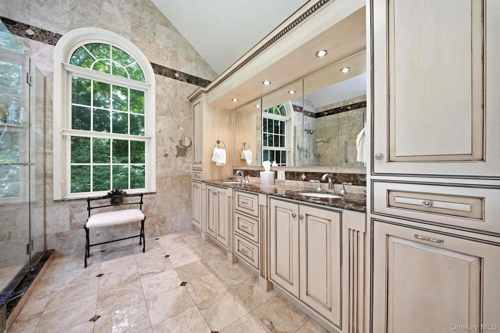 Spacious bathroom featuring a large arched window, extensive vanity cabinetry, dual undermount sinks, and stone tile flooring Spacious bathroom featuring a large arched window, extensive vanity cabinetry, dual undermount sinks, and stone tile flooring