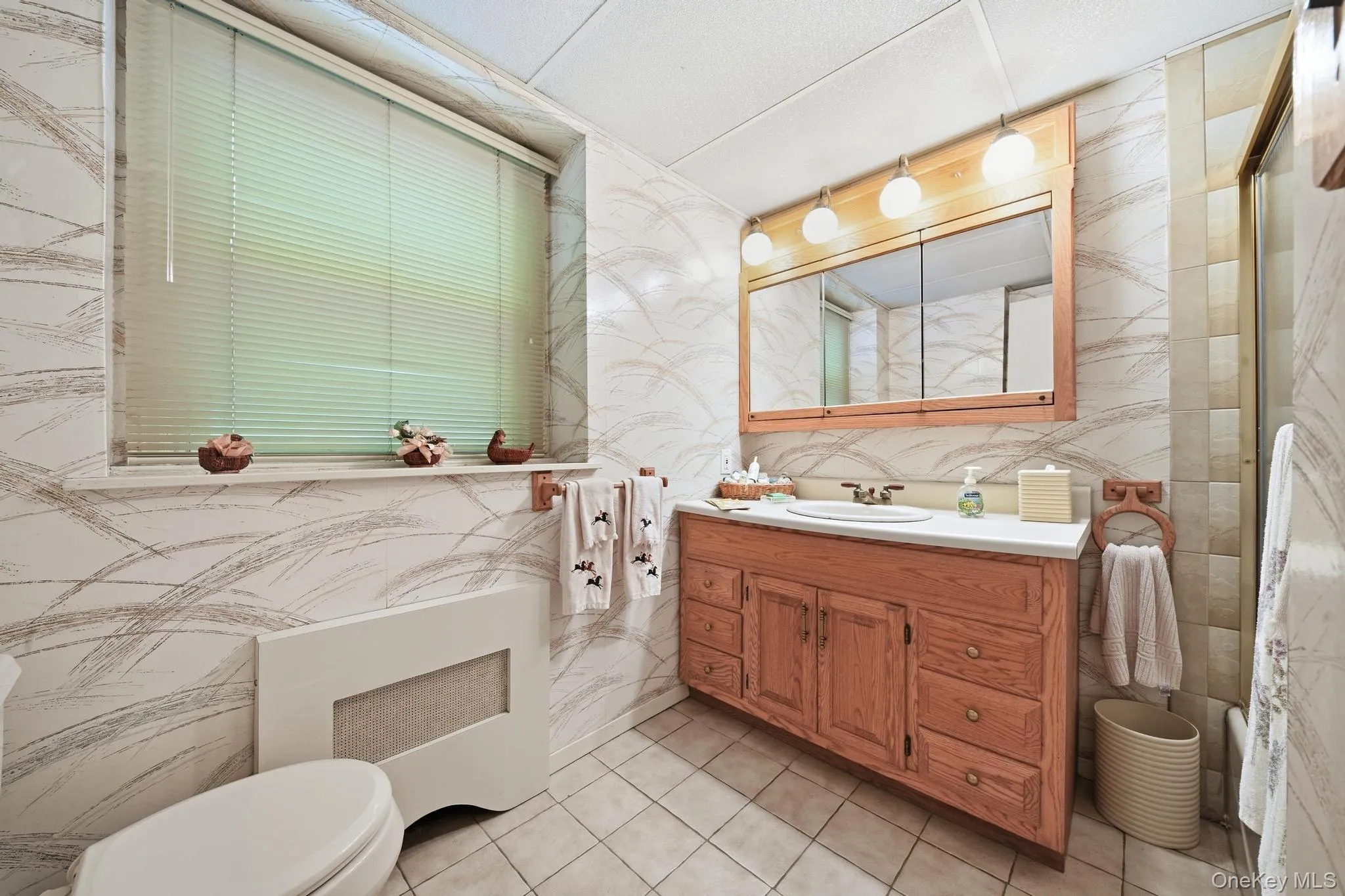 Bathroom featuring a wood-finish vanity with a built-in sink, a wall-mounted medicine cabinet with integrated lighting, tiled flooring, and a window with horizontal blinds Bathroom featuring a wood-finish vanity with a built-in sink, a wall-mounted medicine cabinet with integrated lighting, tiled flooring, and a window with horizontal blinds