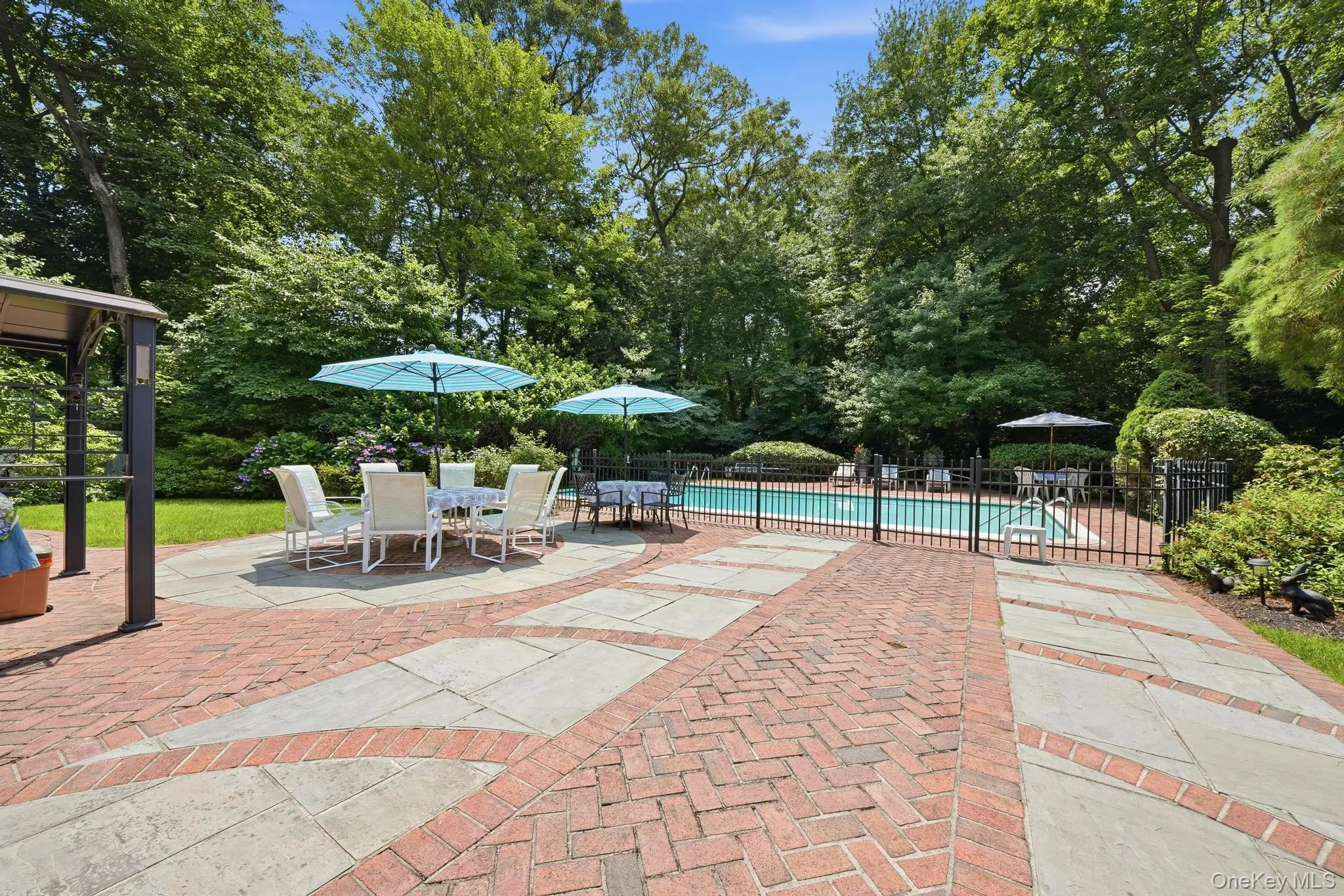 Expansive outdoor living area featuring a brick and stone patio, an in-ground swimming pool with a black metal fence, and mature perimeter trees Expansive outdoor living area featuring a brick and stone patio, an in-ground swimming pool with a black metal fence, and mature perimeter trees