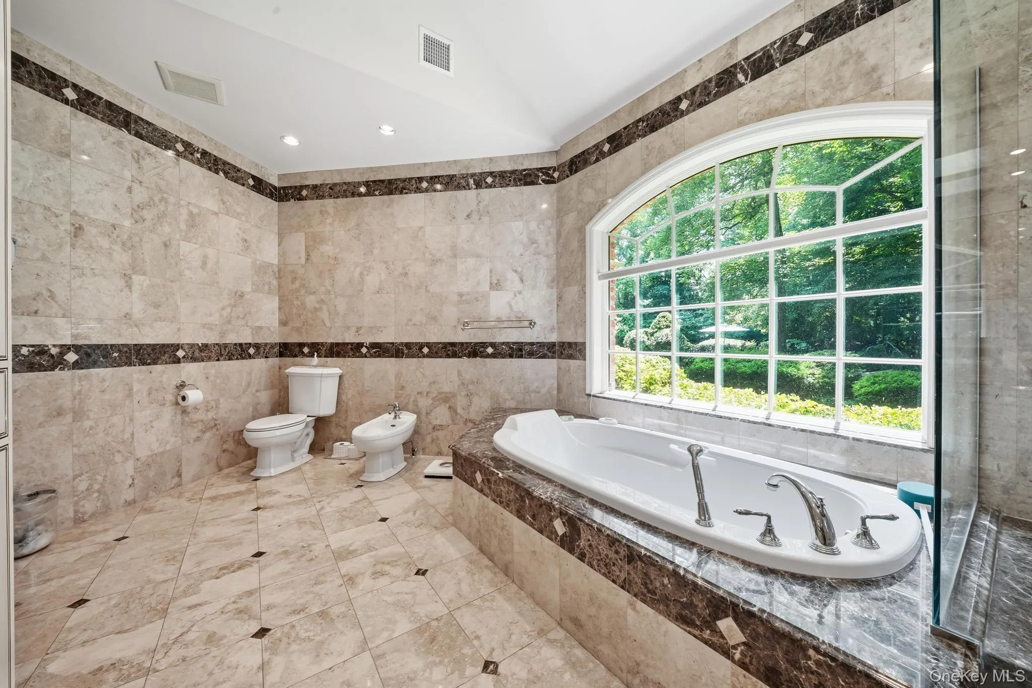 Spacious bathroom featuring an arched window, built-in soaking tub with deck-mounted fixtures, glass-enclosed shower, and full tile surrounds Spacious bathroom featuring an arched window, built-in soaking tub with deck-mounted fixtures, glass-enclosed shower, and full tile surrounds