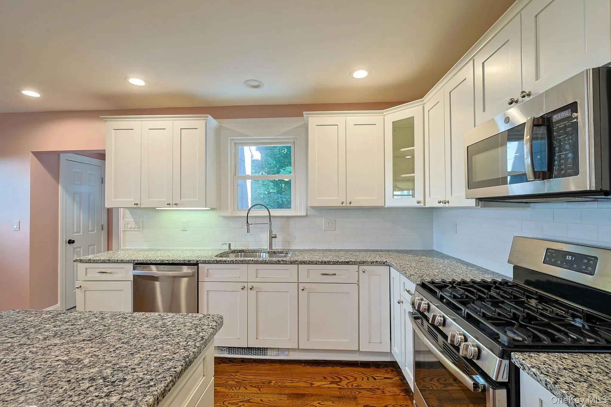 Kitchen featuring white shaker-style cabinetry, granite countertops, subway tile backsplash, stainless steel appliances, and wood-finish flooring Kitchen featuring white shaker-style cabinetry, granite countertops, subway tile backsplash, stainless steel appliances, and wood-finish flooring