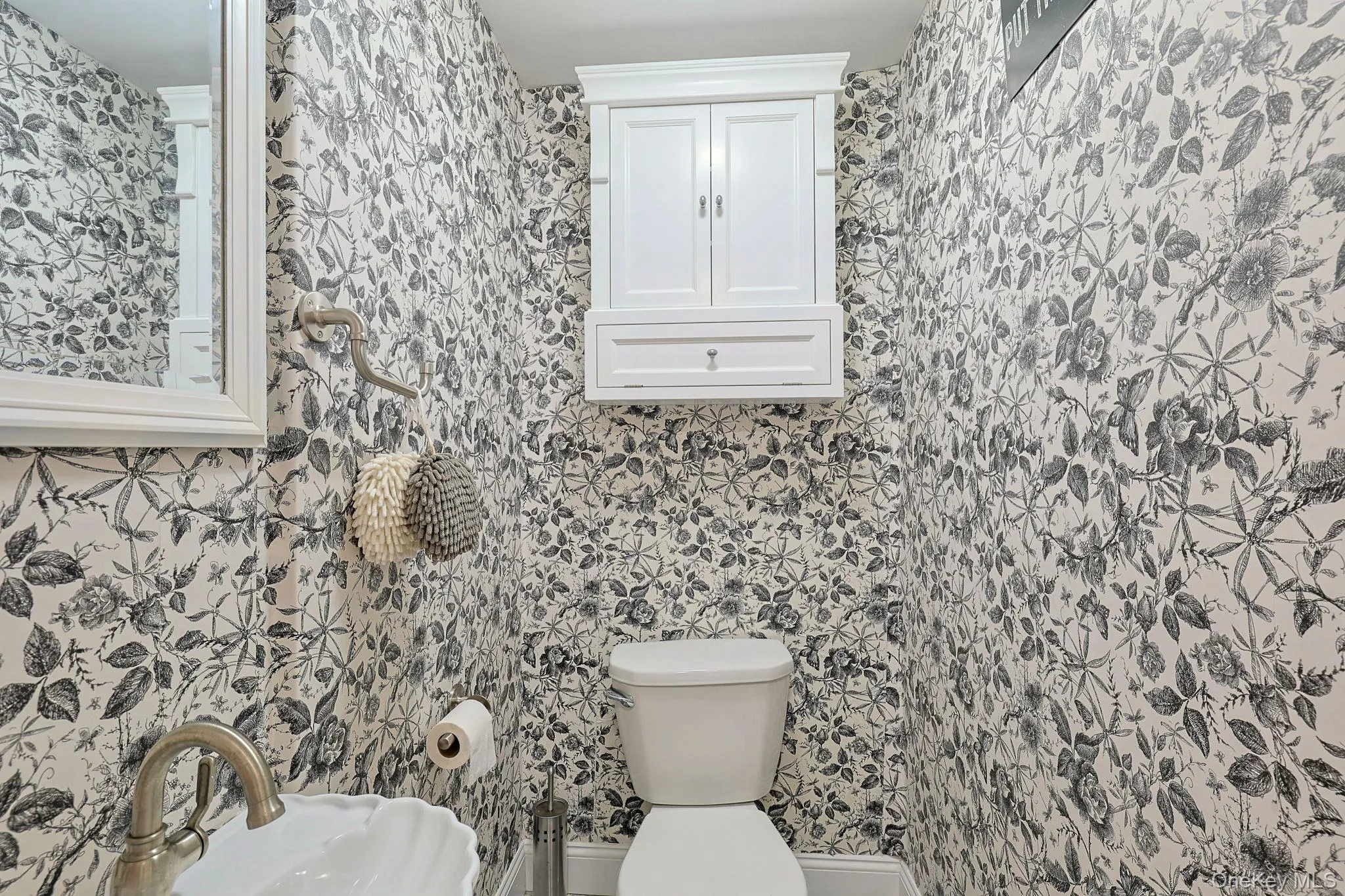 Powder room featuring botanical print wallpaper, a white pedestal toilet, and a wall-mounted white cabinet with paneling Powder room featuring botanical print wallpaper, a white pedestal toilet, and a wall-mounted white cabinet with paneling