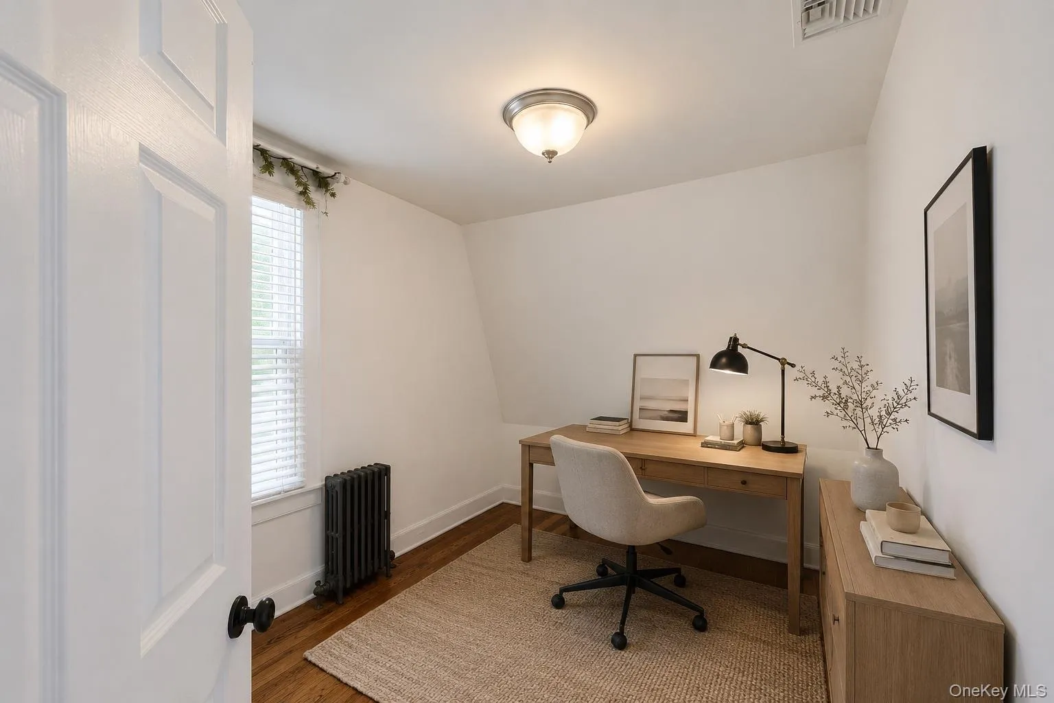 Virtually-staged bedroom as home office. Wood-finish flooring with white baseboards, a window featuring blinds, and a built-in radiator Virtually-staged bedroom as home office. Wood-finish flooring with white baseboards, a window featuring blinds, and a built-in radiator