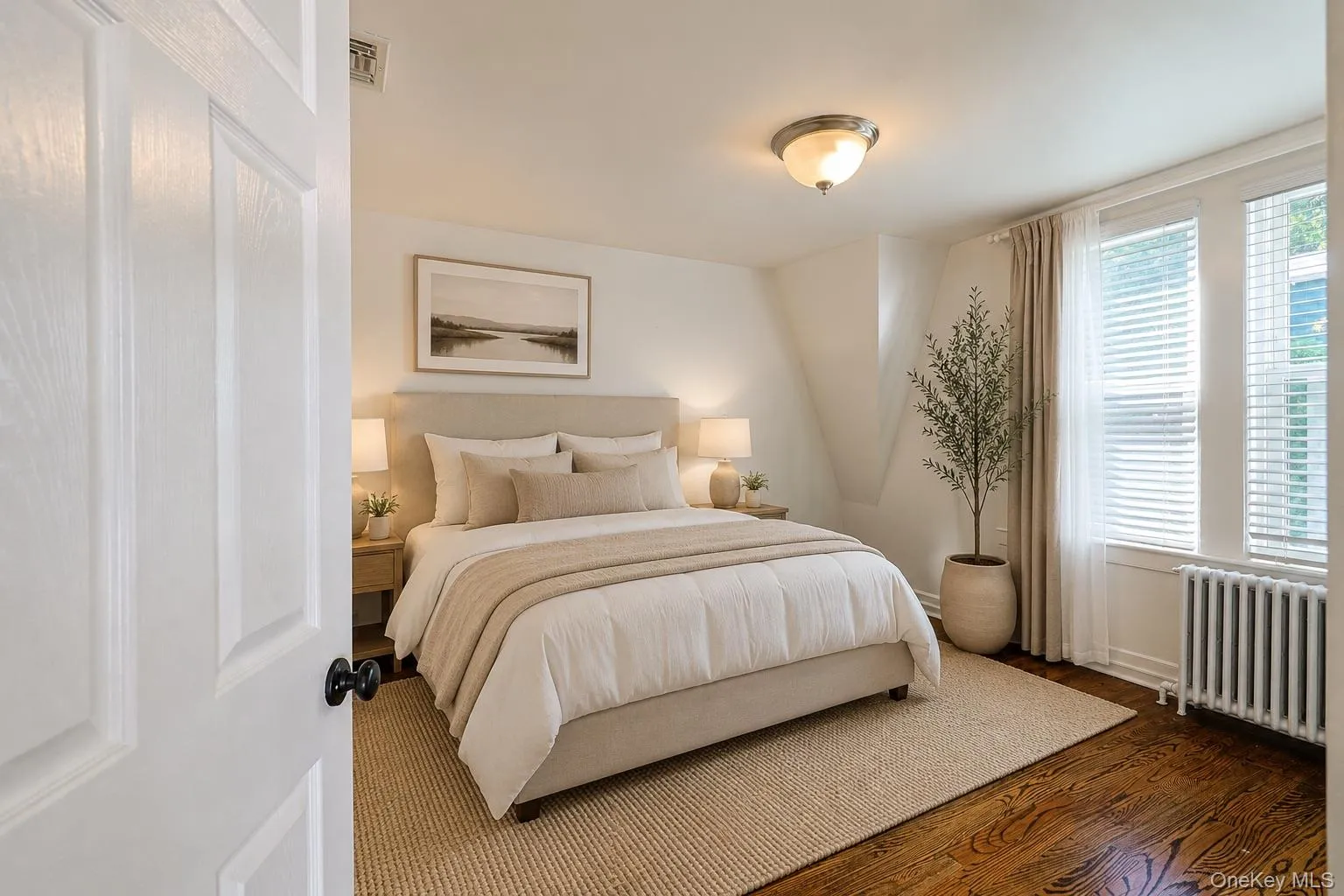 Virtually-staged primary bedroom featuring wood-finish flooring, white paneled door with black hardware, and an overhead dome light fixture Virtually-staged primary bedroom featuring wood-finish flooring, white paneled door with black hardware, and an overhead dome light fixture