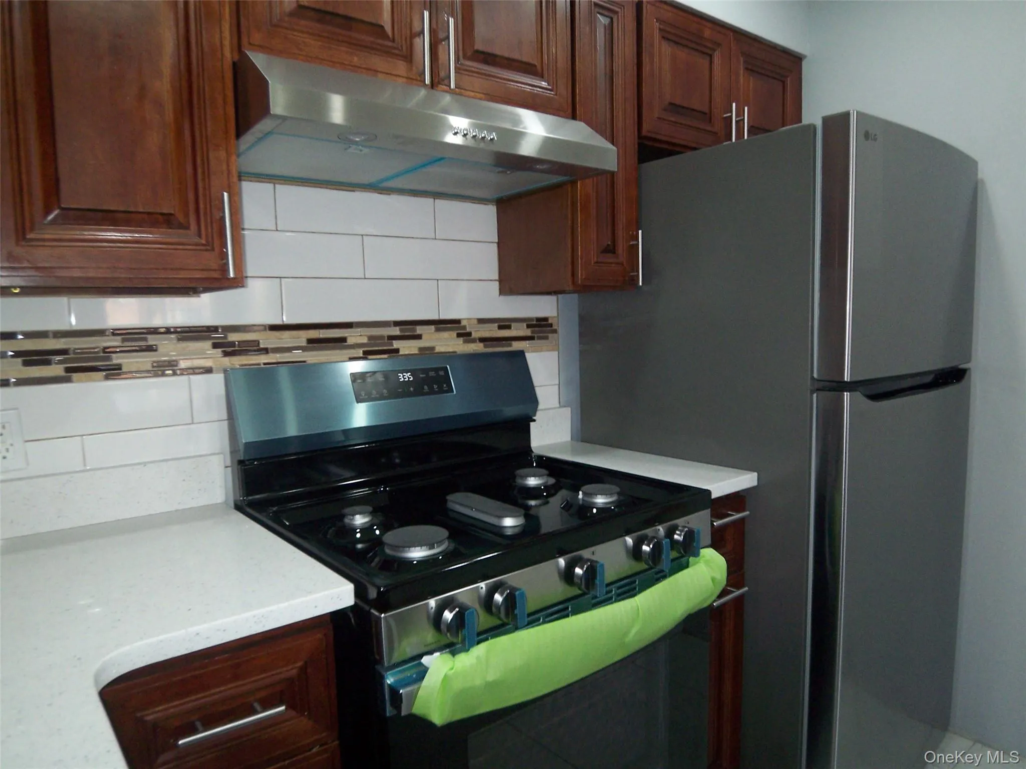 Kitchen featuring stainless steel appliances, wood cabinetry, white subway tile backsplash, decorative mosaic tile accent, and white countertops Kitchen featuring stainless steel appliances, wood cabinetry, white subway tile backsplash, decorative mosaic tile accent, and white countertops
