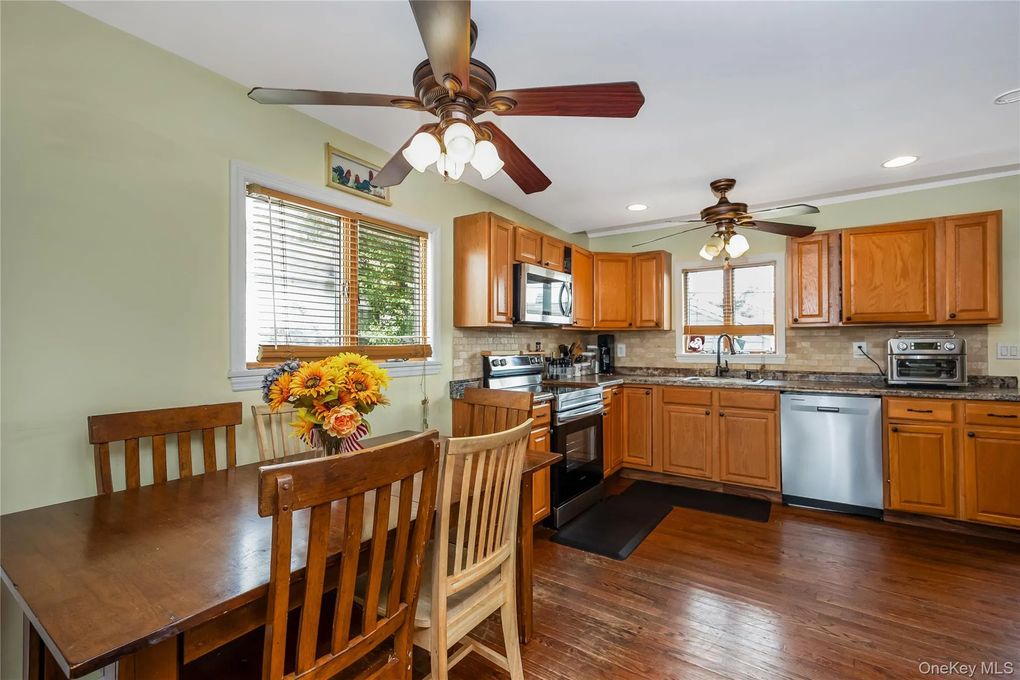 Kitchen and dining area featuring wood-finish flooring, wood cabinetry, stainless steel appliances, and ceiling fans with integrated lighting Kitchen and dining area featuring wood-finish flooring, wood cabinetry, stainless steel appliances, and ceiling fans with integrated lighting