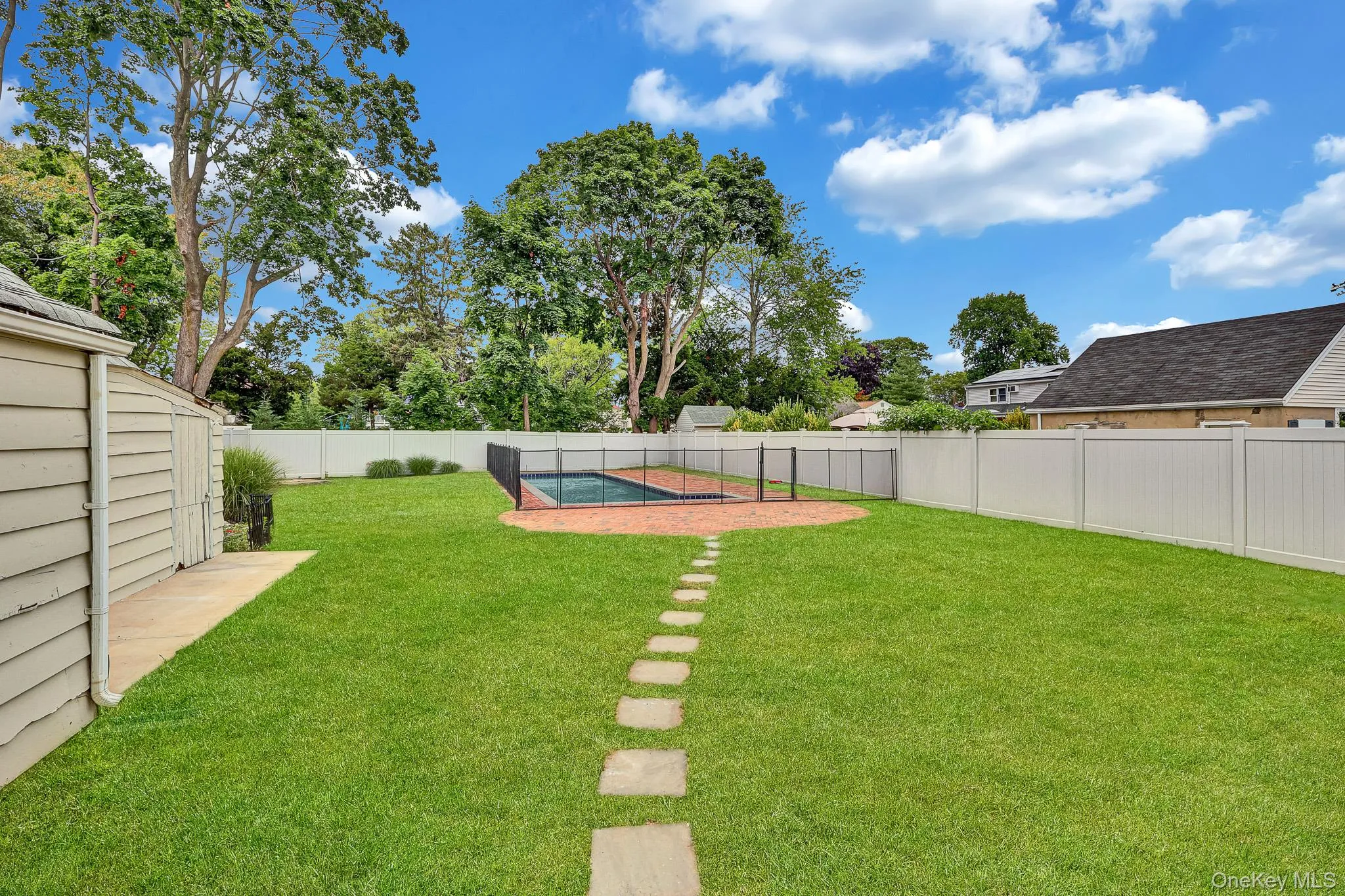 Expansive backyard featuring an in-ground swimming pool with safety fencing, a stone paver path, a white privacy fence, and a storage shed Expansive backyard featuring an in-ground swimming pool with safety fencing, a stone paver path, a white privacy fence, and a storage shed