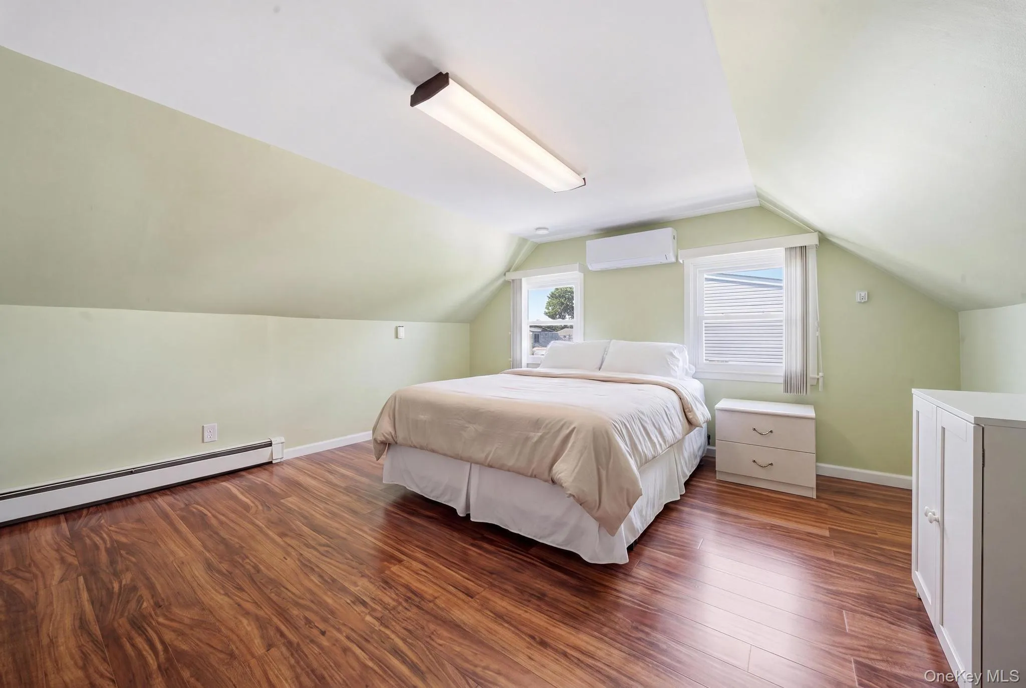 Attic room featuring rich wood-finish flooring, light green walls, two windows with white blinds, and a wall-mounted heating/cooling unit Attic room featuring rich wood-finish flooring, light green walls, two windows with white blinds, and a wall-mounted heating/cooling unit