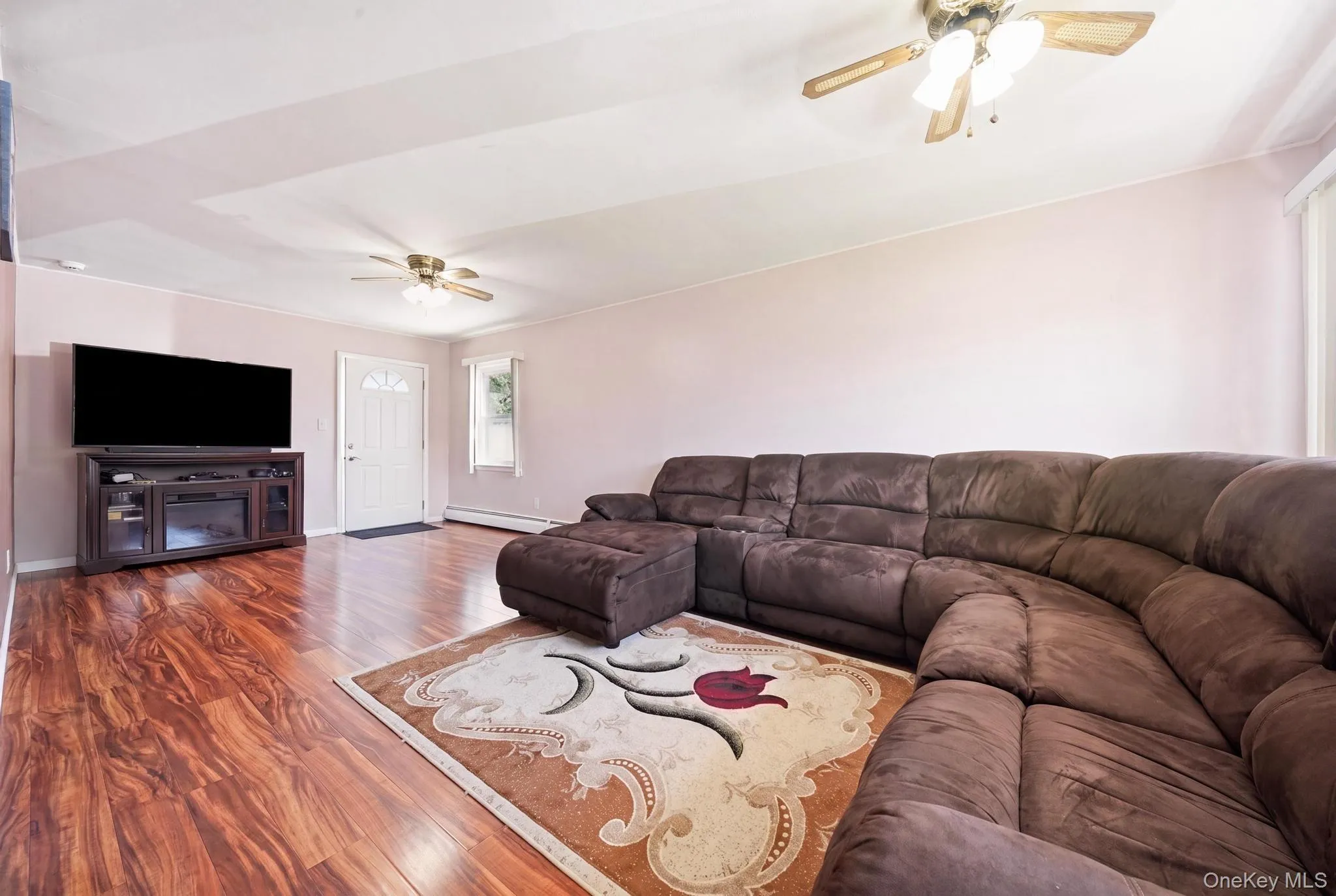 Spacious living area featuring wood-finish flooring, a ceiling fan, and a white entry door with frosted glass accents Spacious living area featuring wood-finish flooring, a ceiling fan, and a white entry door with frosted glass accents