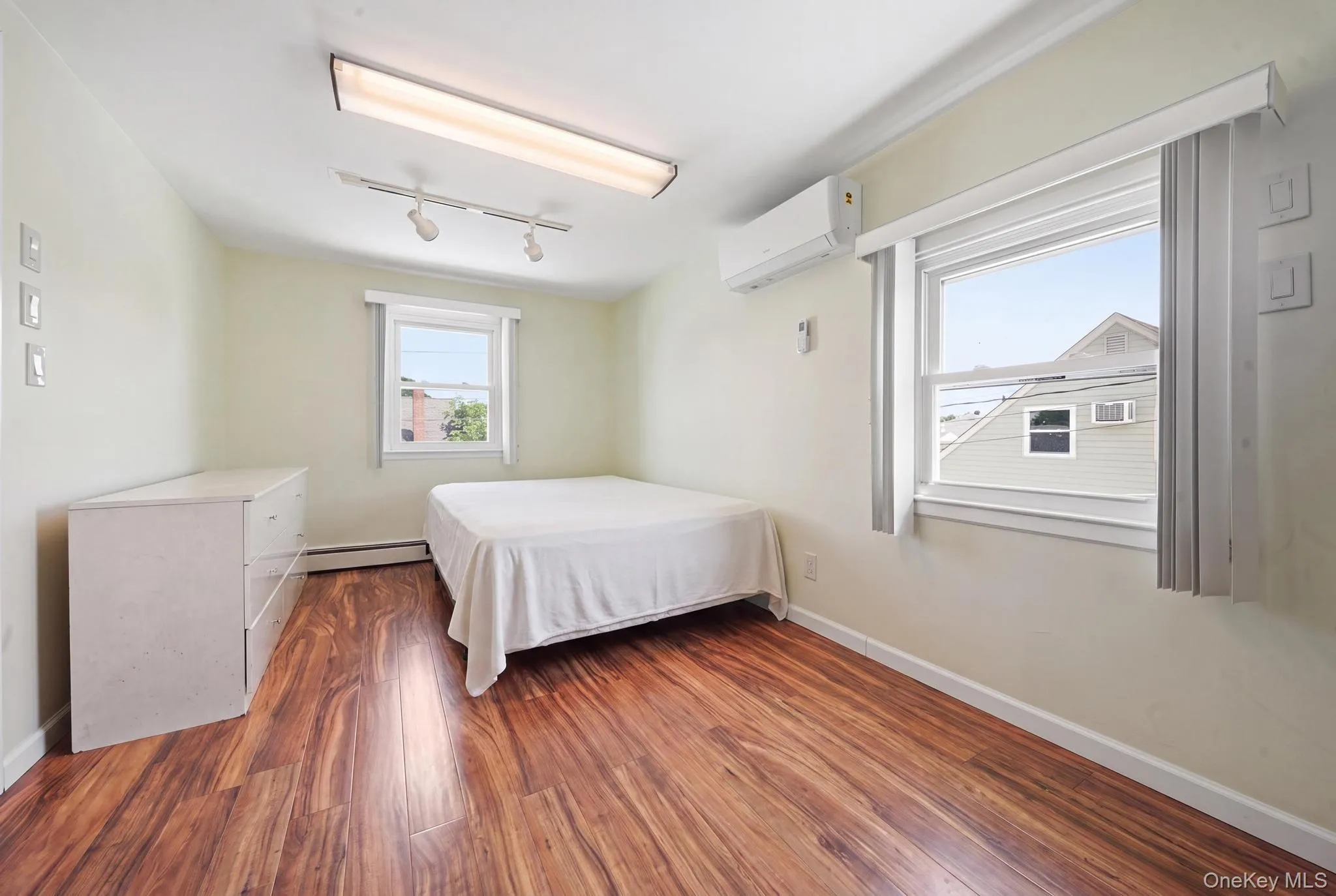 Room featuring wood-finish flooring, two windows, baseboard heating, a ceiling-mounted fluorescent light fixture, and a wall-mounted mini-split system Room featuring wood-finish flooring, two windows, baseboard heating, a ceiling-mounted fluorescent light fixture, and a wall-mounted mini-split system