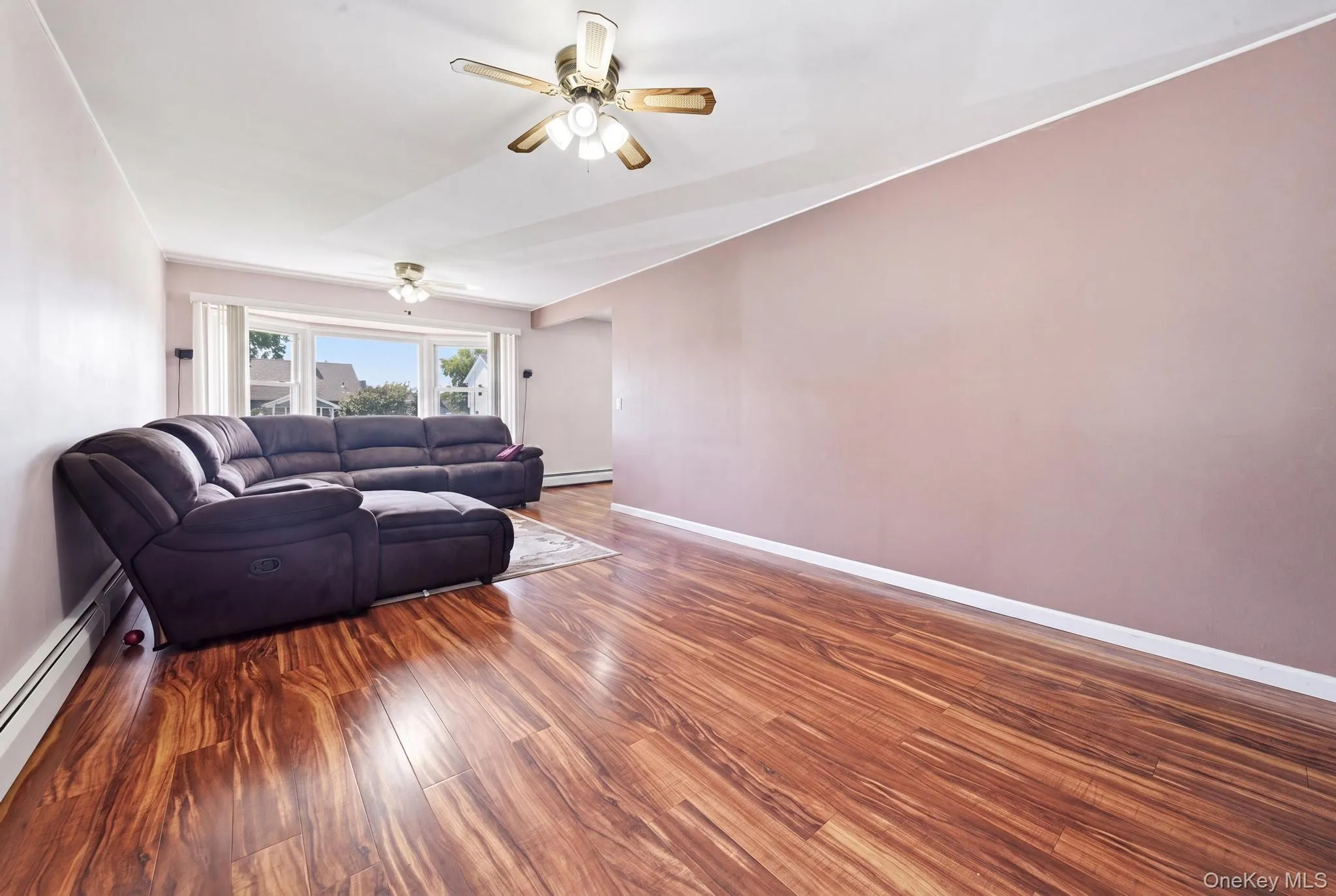 Spacious interior featuring wood-finish flooring, a ceiling fan with integrated lighting, and a bay window providing natural illumination Spacious interior featuring wood-finish flooring, a ceiling fan with integrated lighting, and a bay window providing natural illumination