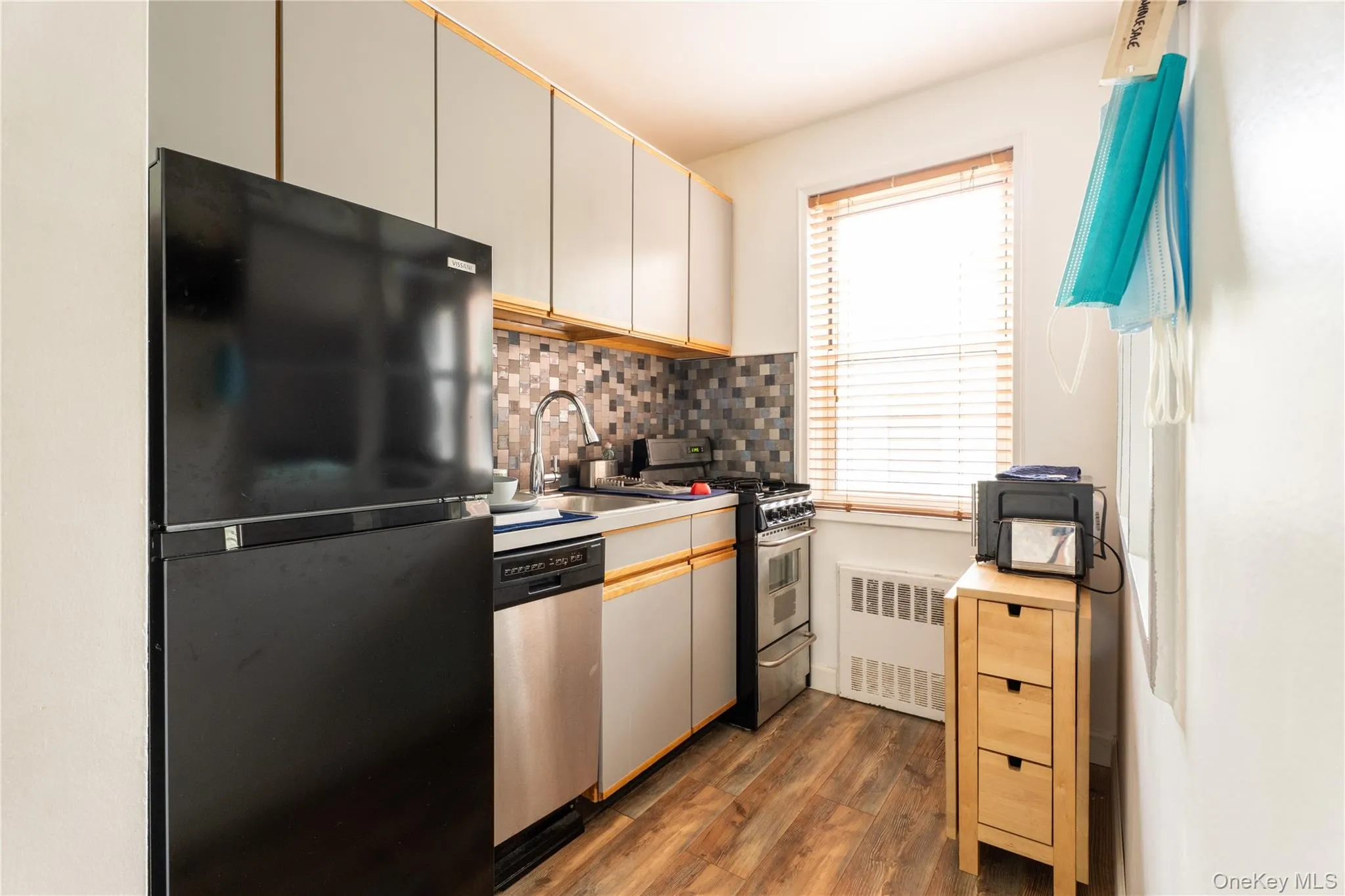 Compact kitchen featuring wood-finish flooring, a mosaic tile backsplash, a stainless steel dishwasher, a gas range, and a window with blinds Compact kitchen featuring wood-finish flooring, a mosaic tile backsplash, a stainless steel dishwasher, a gas range, and a window with blinds
