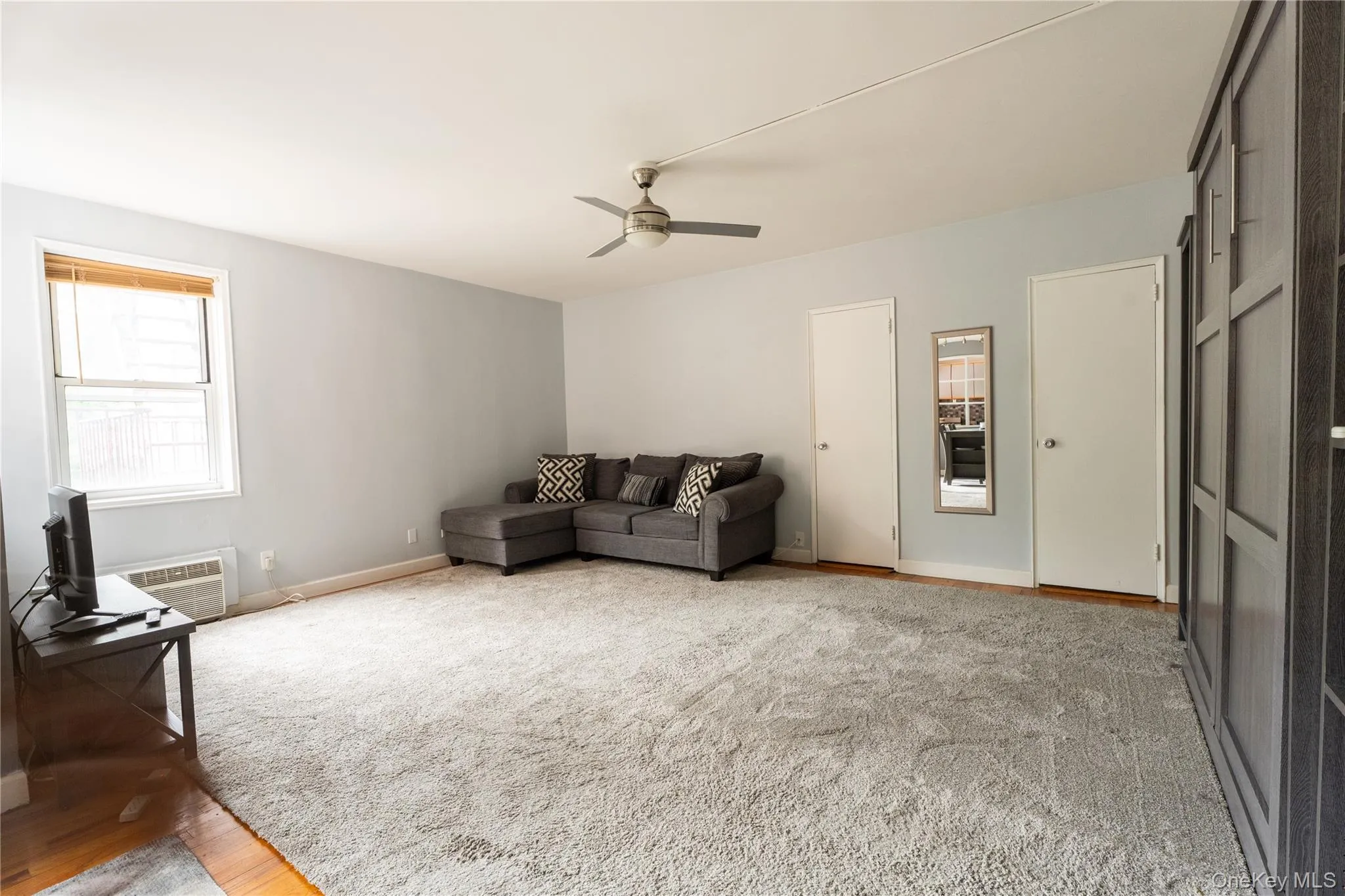Spacious living area featuring light gray wall paint, light-colored carpeting, a window with horizontal blinds, a ceiling fan, and built-in shelving units Spacious living area featuring light gray wall paint, light-colored carpeting, a window with horizontal blinds, a ceiling fan, and built-in shelving units