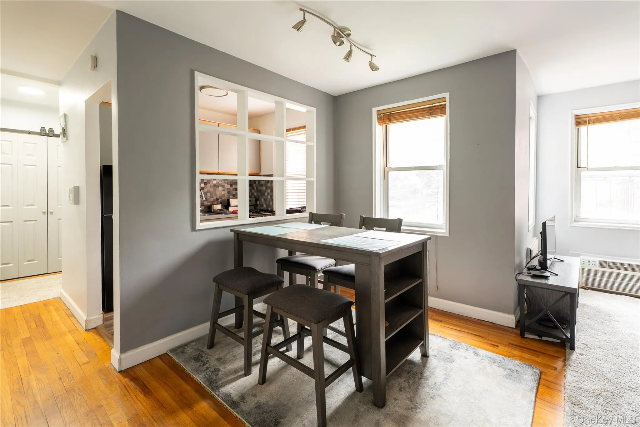 Living area featuring a pass-through window to the kitchen, wood-finish flooring, two windows with blinds, and track lighting Living area featuring a pass-through window to the kitchen, wood-finish flooring, two windows with blinds, and track lighting