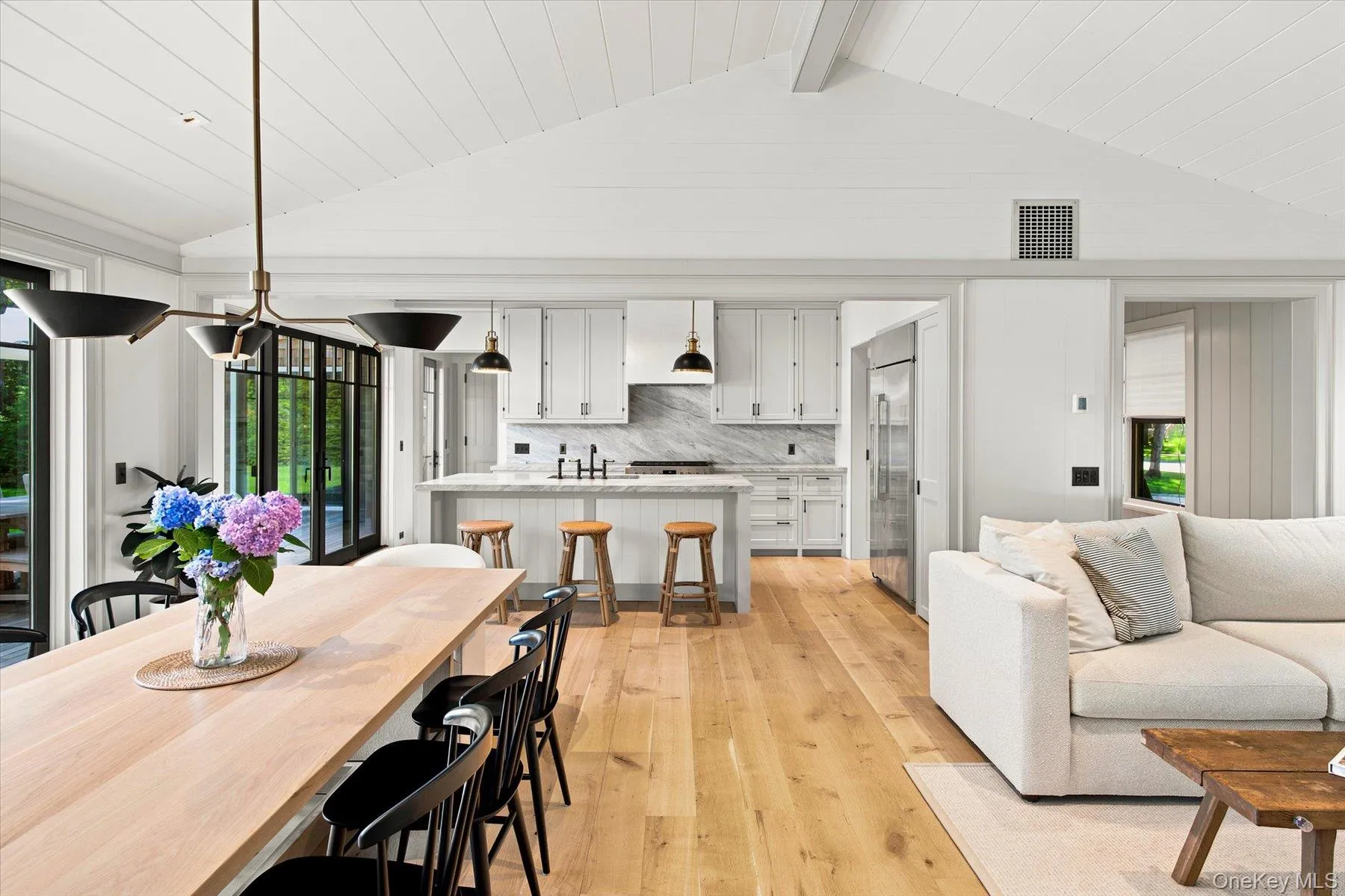 Vaulted ceiling with exposed beams, light wood-finish flooring, full-height black-framed glass doors, white kitchen cabinetry with integrated appliances, and a kitchen island featuring a waterfall countertop Vaulted ceiling with exposed beams, light wood-finish flooring, full-height black-framed glass doors, white kitchen cabinetry with integrated appliances, and a kitchen island featuring a waterfall countertop