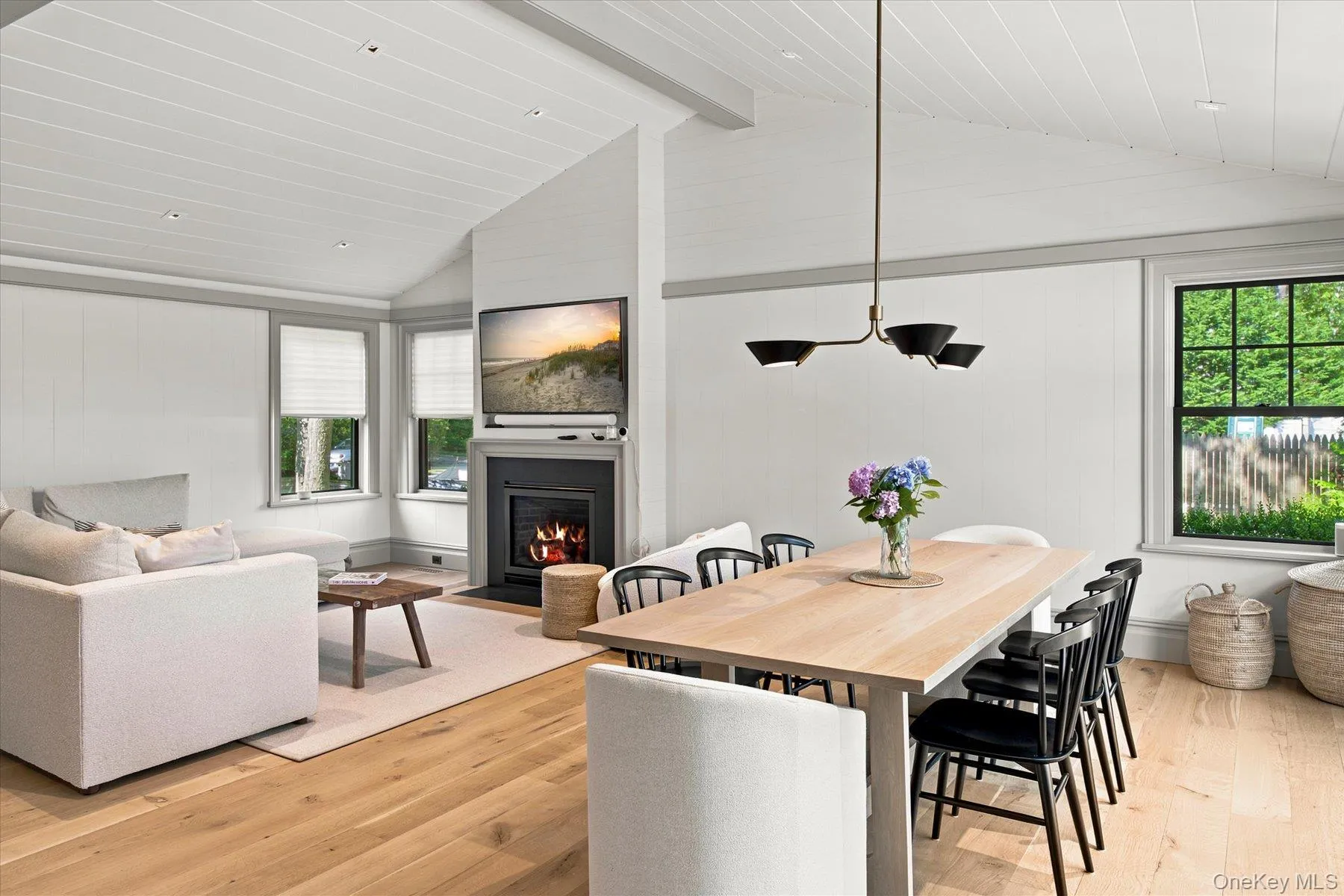 Open-concept living and dining area featuring wood-finish flooring, a fireplace with a mounted television, a vaulted ceiling with recessed lighting, and large windows with privacy blinds Open-concept living and dining area featuring wood-finish flooring, a fireplace with a mounted television, a vaulted ceiling with recessed lighting, and large windows with privacy blinds