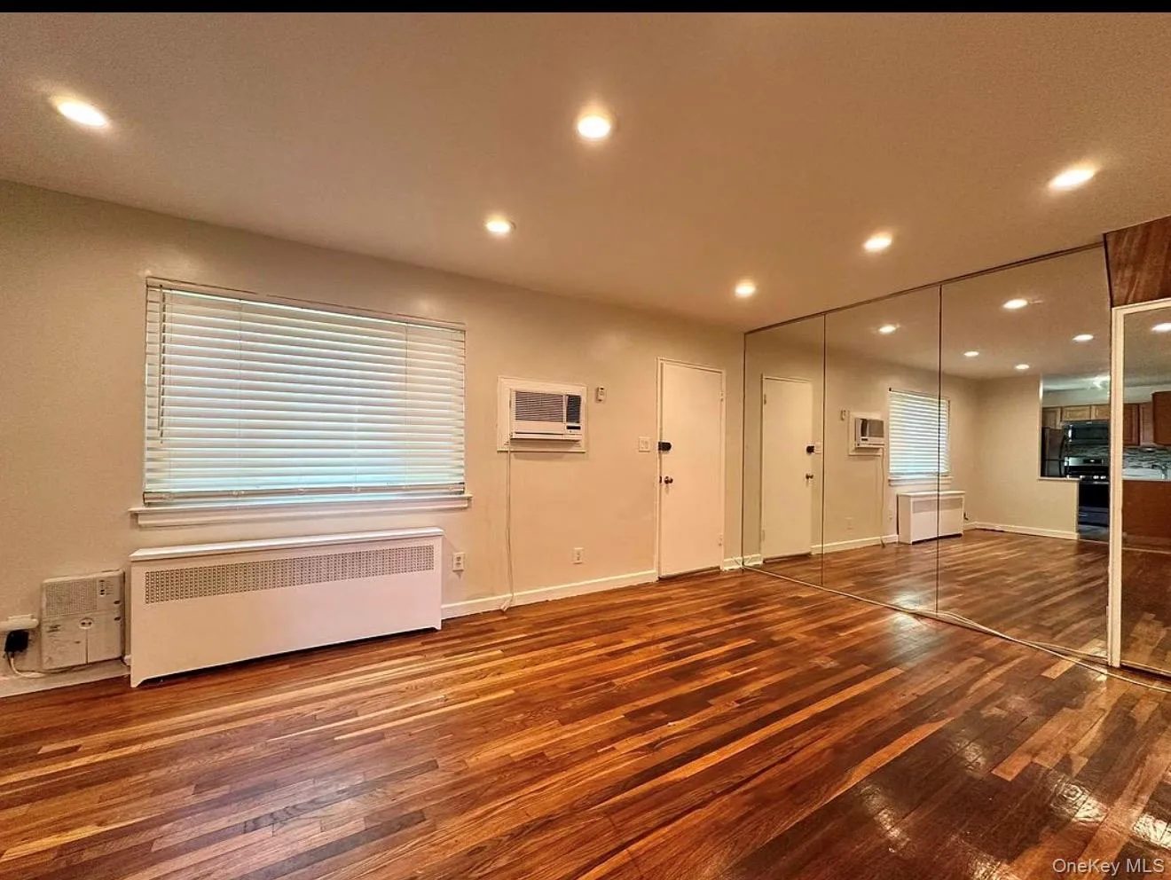 Refinished wood flooring, recessed lighting, expansive mirrored wall, window with blinds, and through-wall air conditioning unit Refinished wood flooring, recessed lighting, expansive mirrored wall, window with blinds, and through-wall air conditioning unit
