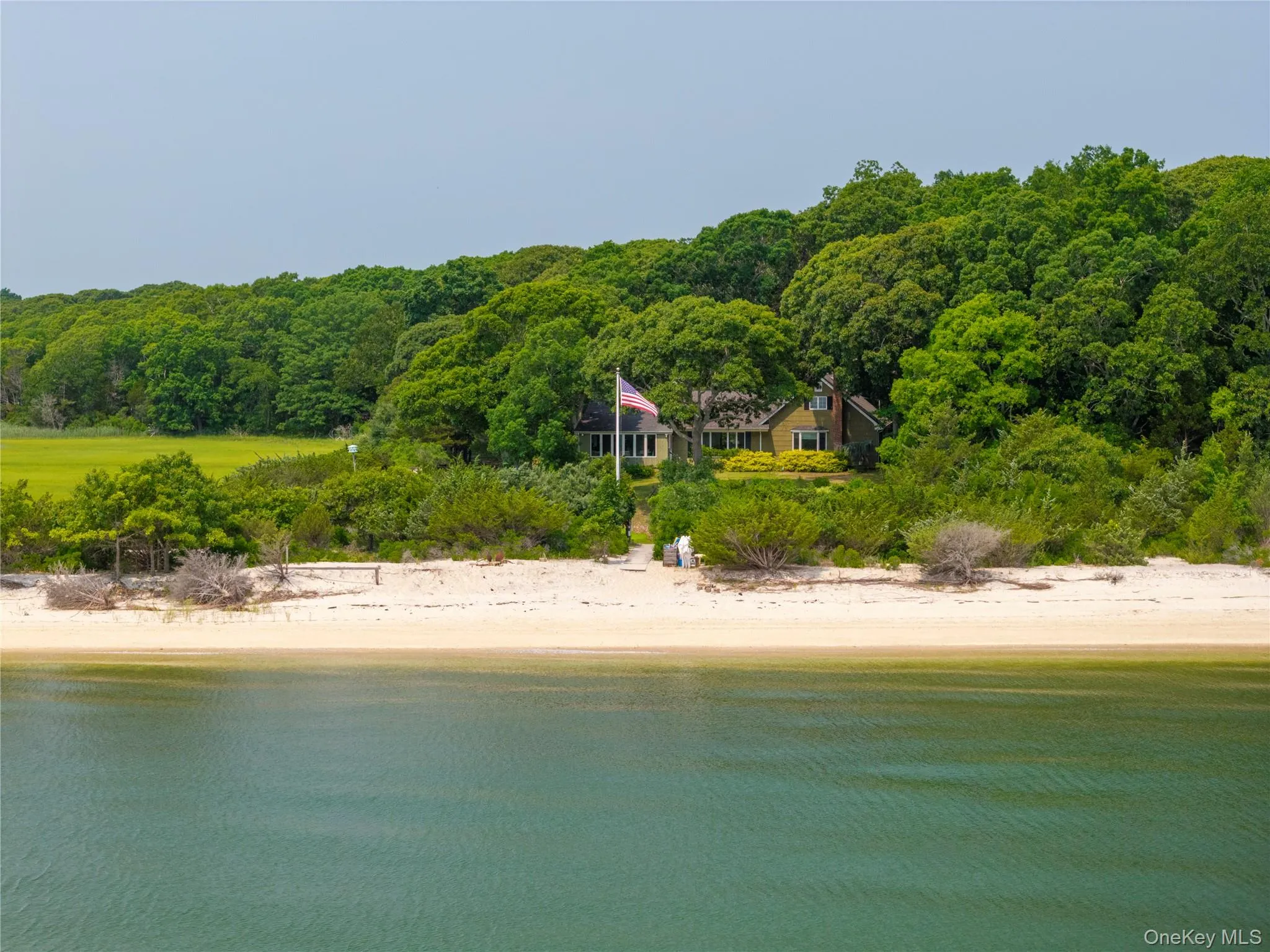 Waterfront property featuring a sandy beach, mature tree line, and a single-family residence with a light-toned exterior and dark roof Waterfront property featuring a sandy beach, mature tree line, and a single-family residence with a light-toned exterior and dark roof
