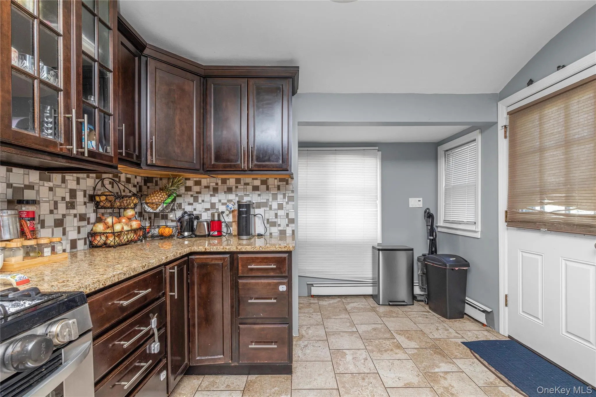 Kitchen featuring dark brown cabinetry, light stone counters, decorative backsplash, a baseboard radiator, and glass insert cabinets Kitchen featuring dark brown cabinetry, light stone counters, decorative backsplash, a baseboard radiator, and glass insert cabinets