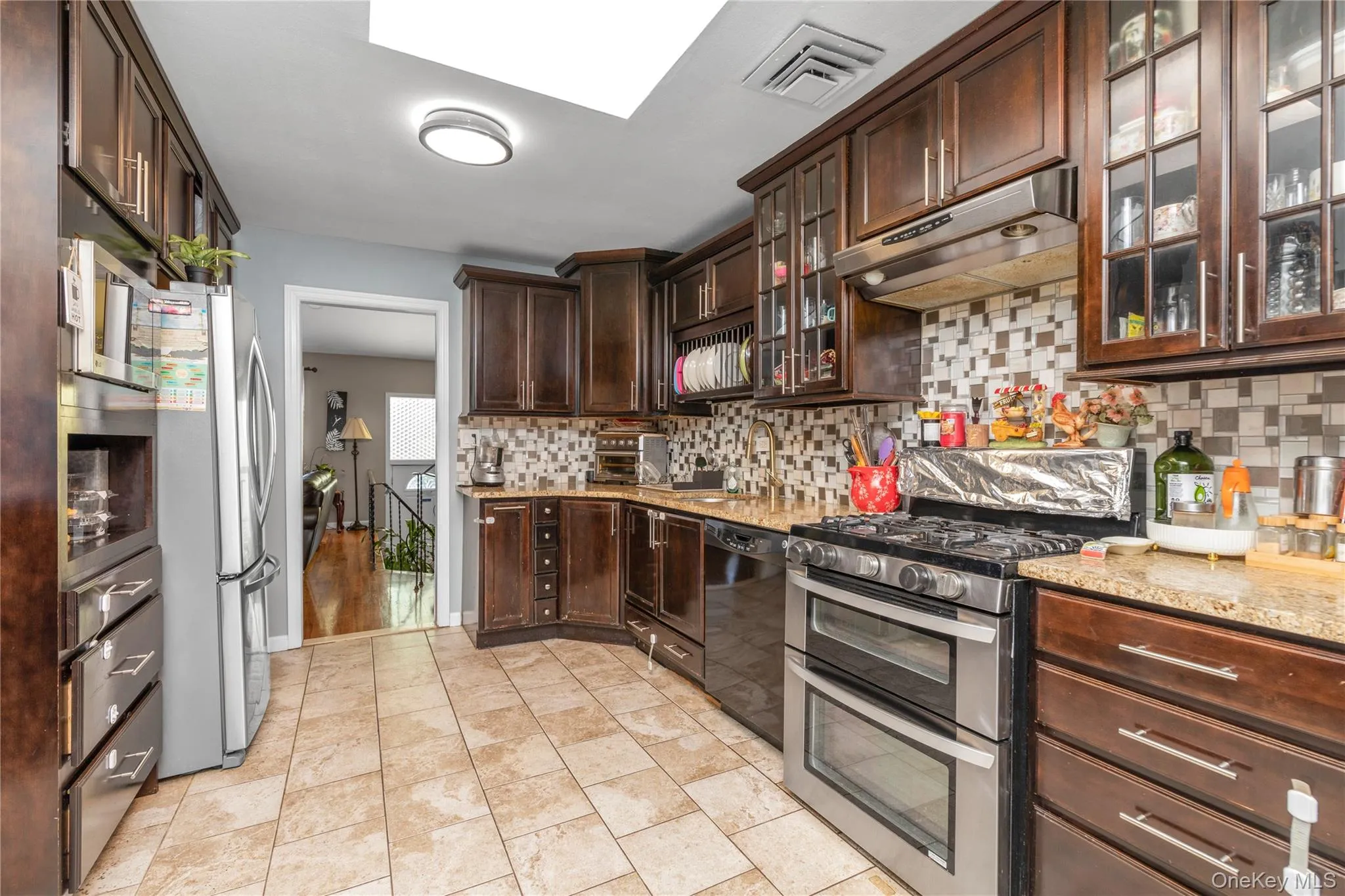 Kitchen featuring backsplash, stainless steel appliances, dark brown cabinetry, under cabinet range hood, and glass insert cabinets Kitchen featuring backsplash, stainless steel appliances, dark brown cabinetry, under cabinet range hood, and glass insert cabinets
