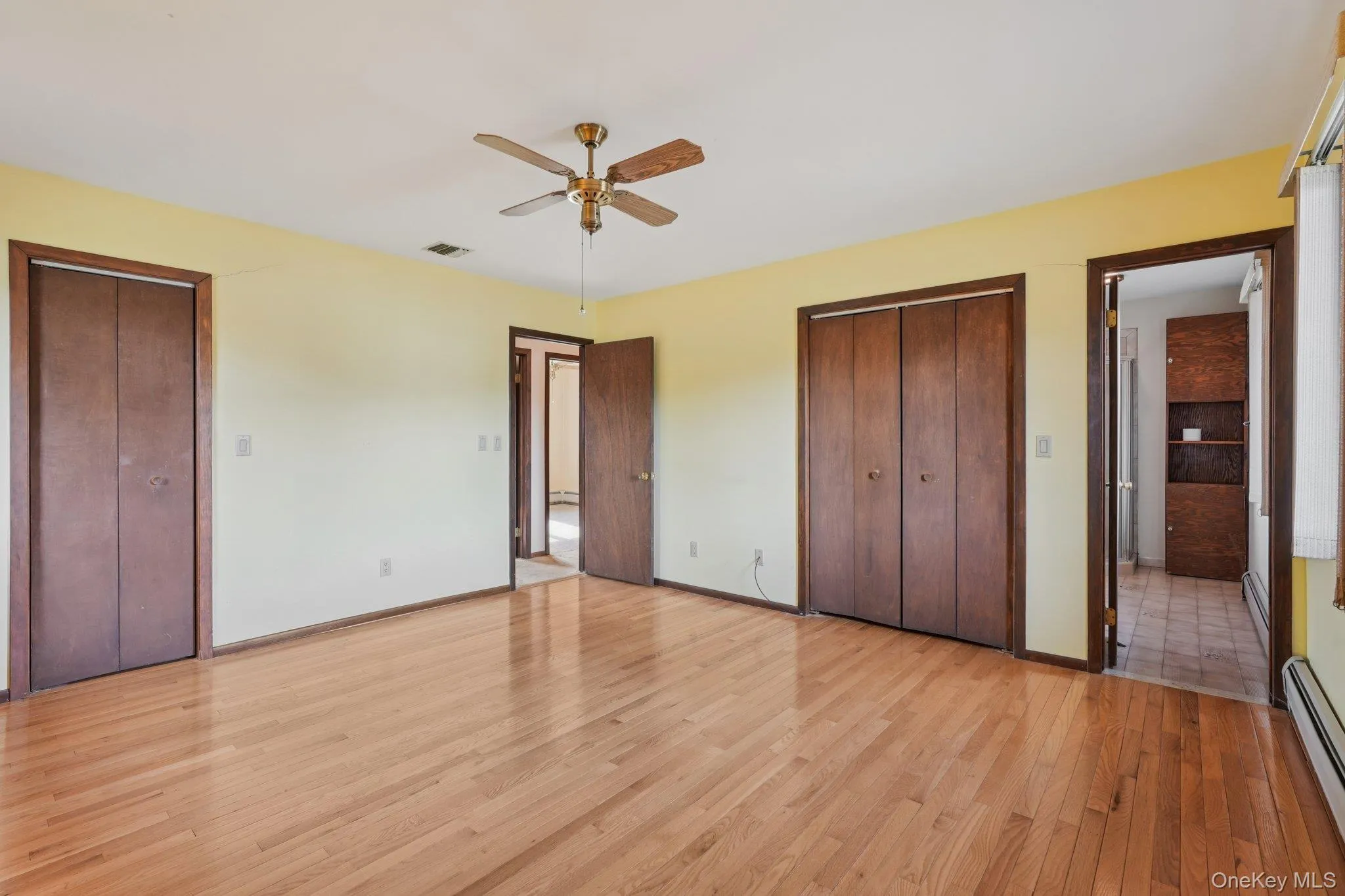 Unfurnished bedroom with two closets, light wood-style flooring, baseboard heating, and a ceiling fan Unfurnished bedroom with two closets, light wood-style flooring, baseboard heating, and a ceiling fan