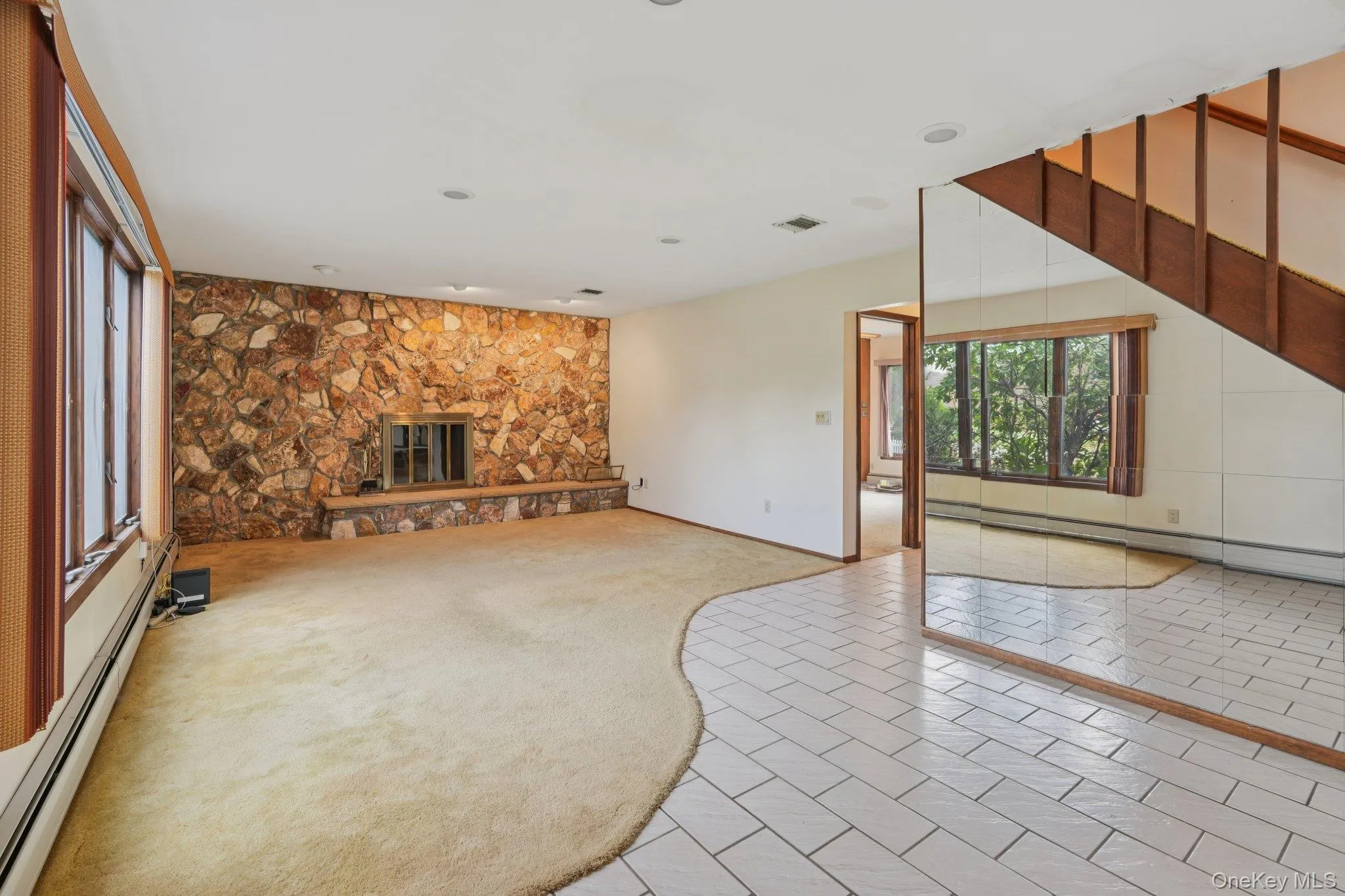 Unfurnished living room with a baseboard heating unit, light tile patterned floors, light carpet, and a stone fireplace Unfurnished living room with a baseboard heating unit, light tile patterned floors, light carpet, and a stone fireplace